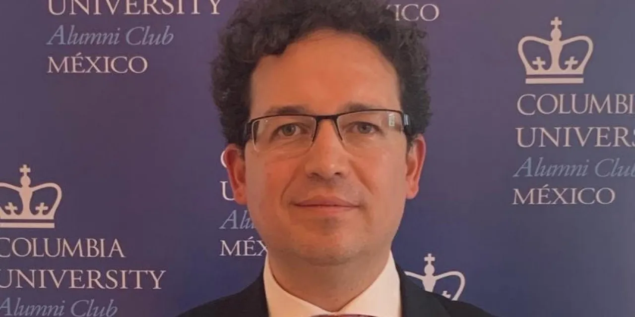 A man with glasses and curly hair poses in front of a backdrop that reads Columbia University Alumni Club Mexico.