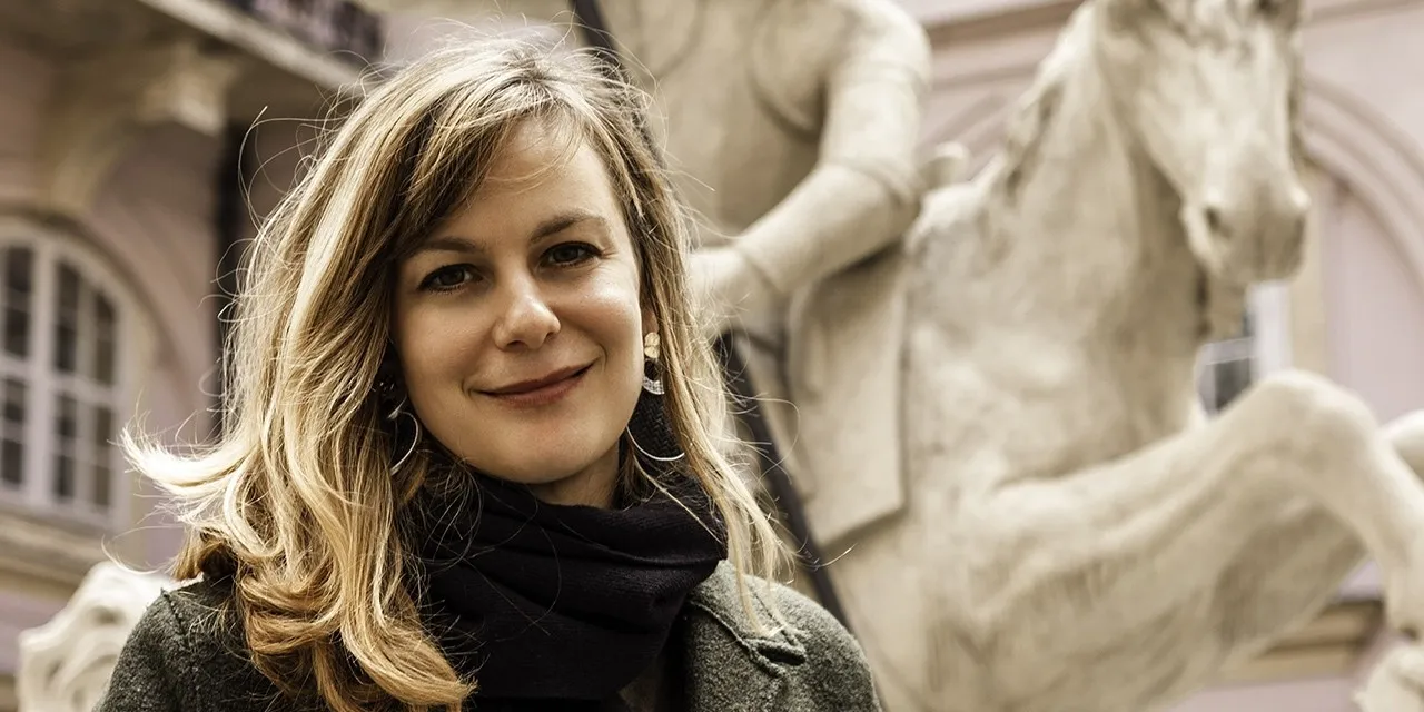 A blonde woman smiles in front of an old stone statue of a horse.