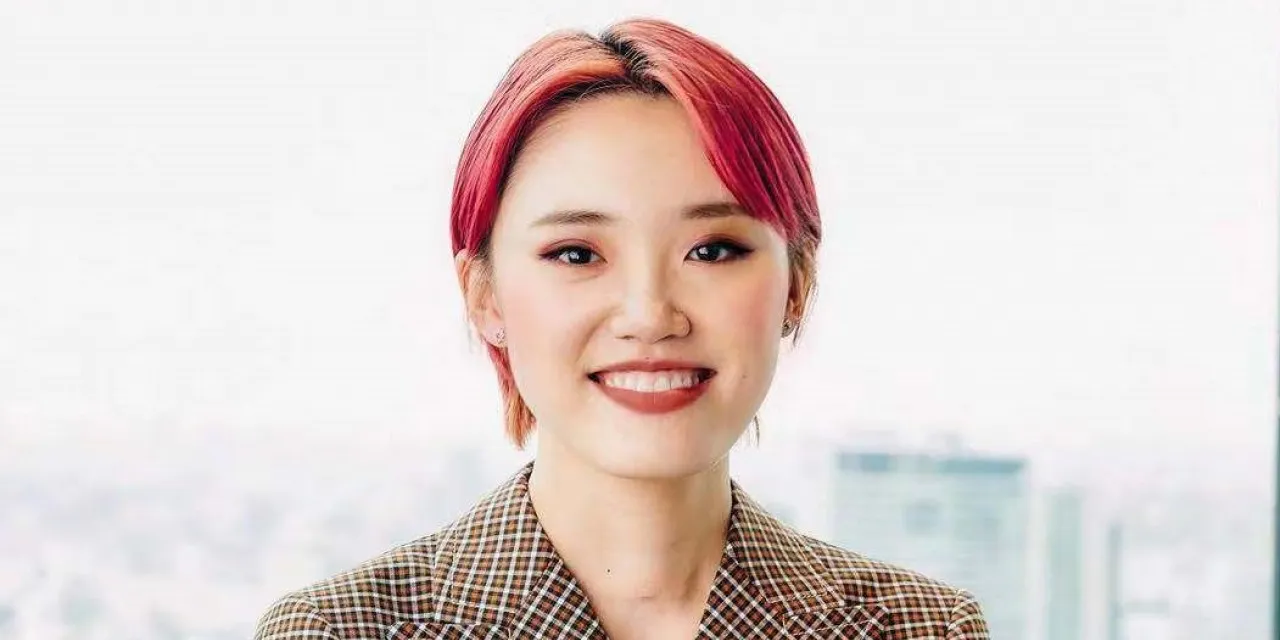 A woman with pink hair wears a checked blazer in front of a city backdrop.