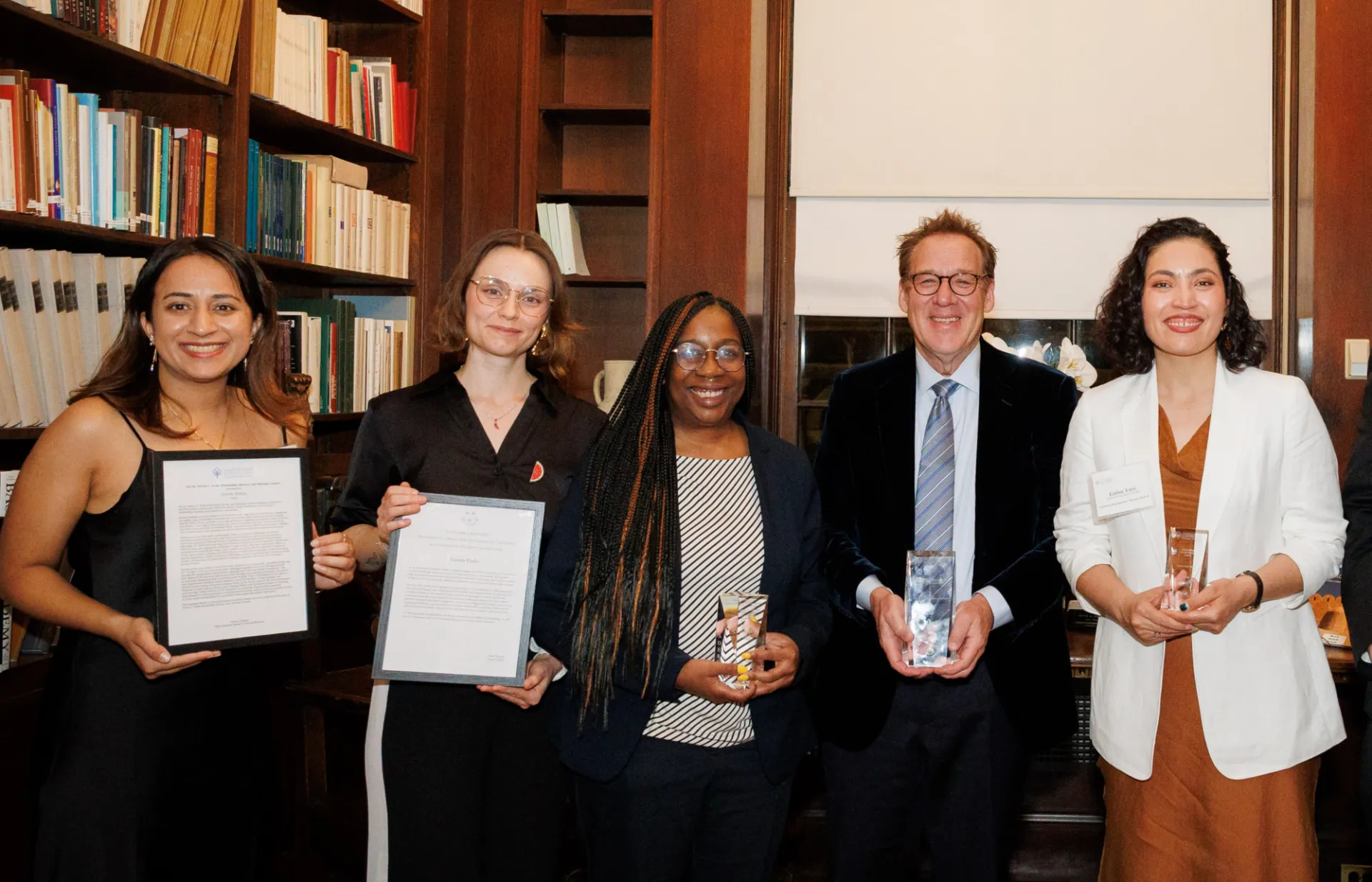A group of people stand smiling in front of bookshelves, each holding an award or certificate. The setting is warm and formal, conveying achievement.