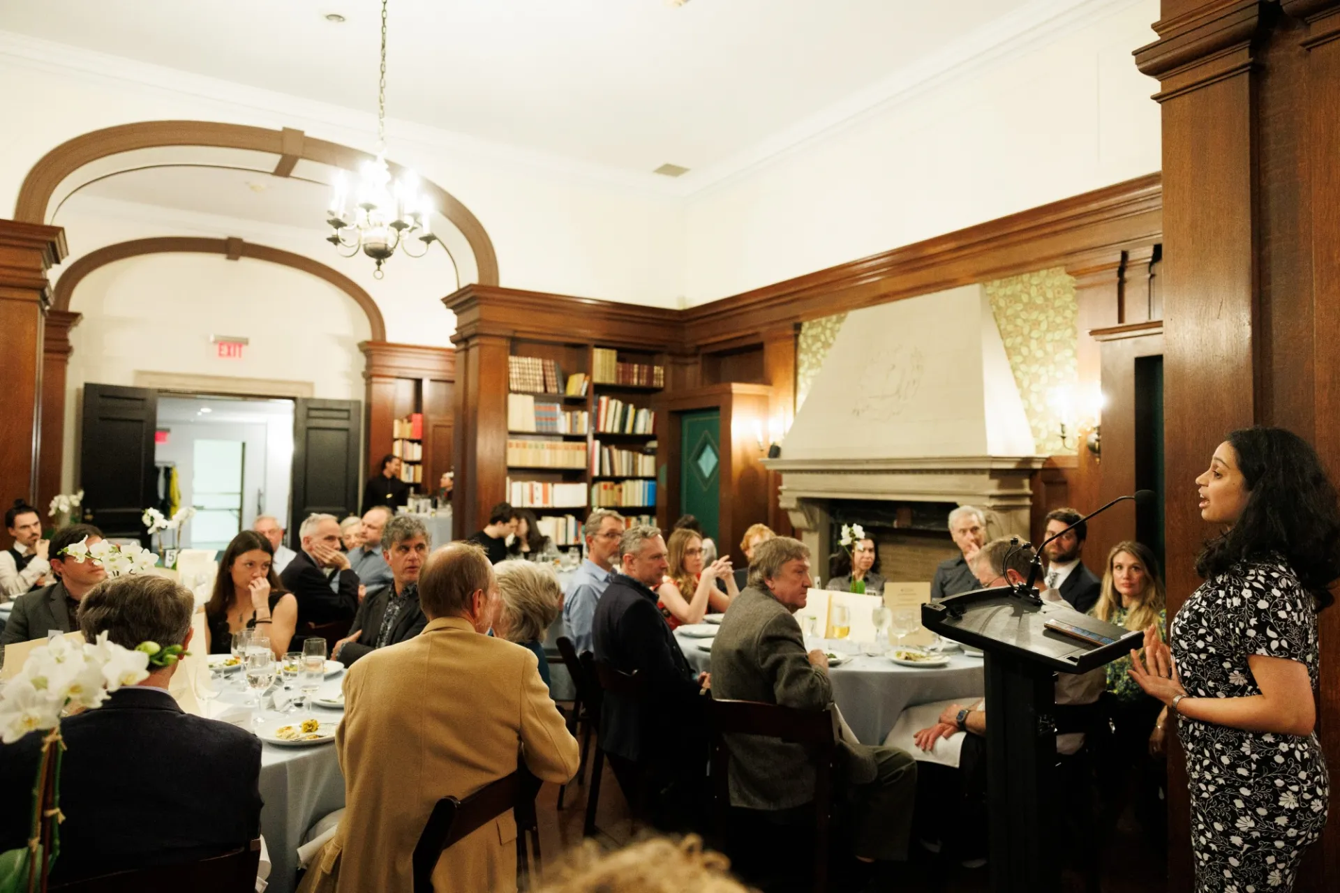 Woman at lectern speaking while people seated at tables listen. 