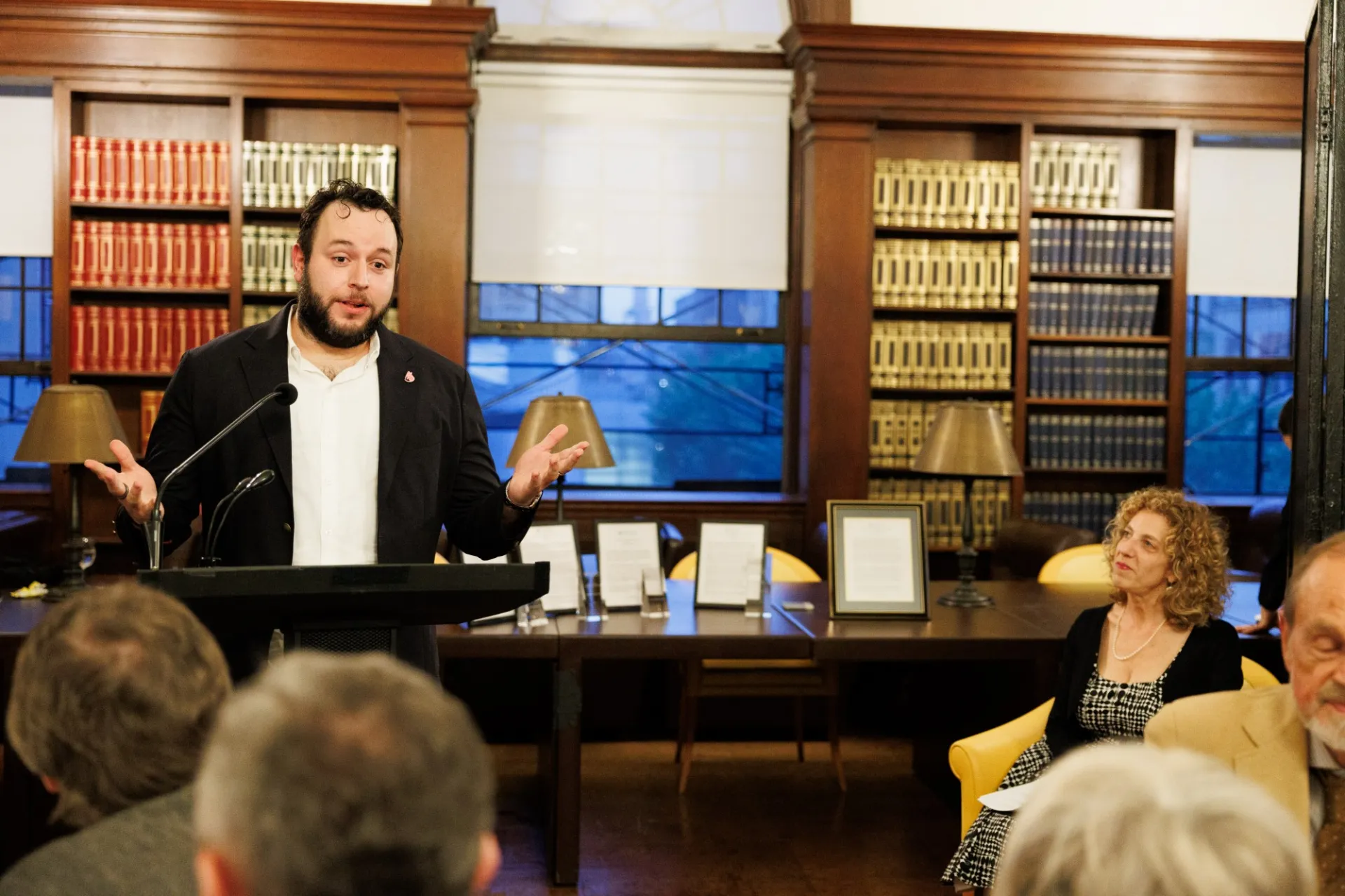 Man at lectern speaking as those seated at tables listen.