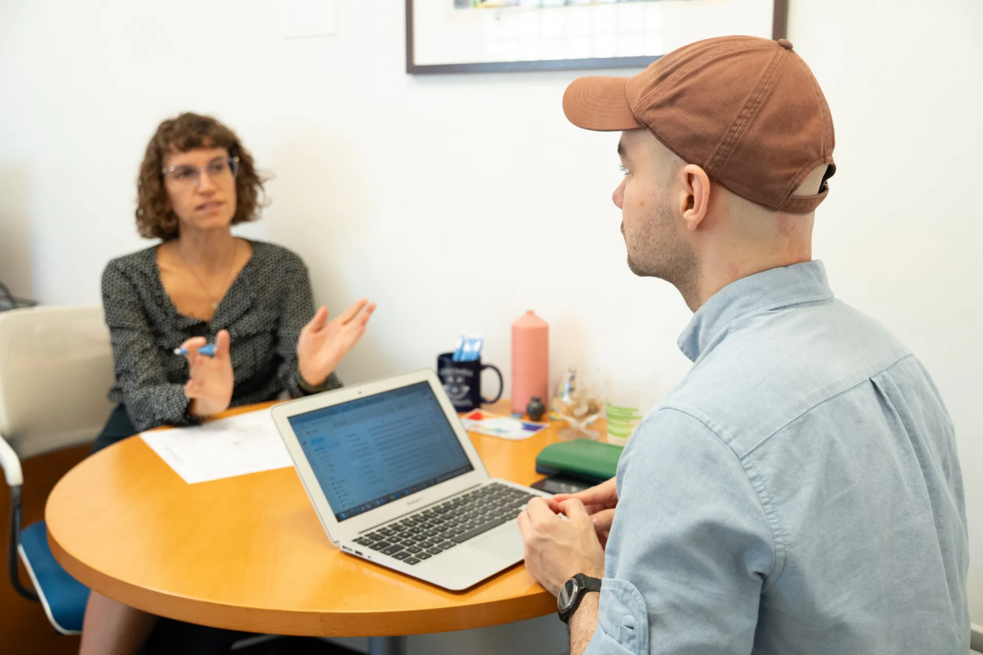 Two people having a discussion at a round table. A woman with curly hair gestures while speaking, and a man in a cap listens, using a laptop.