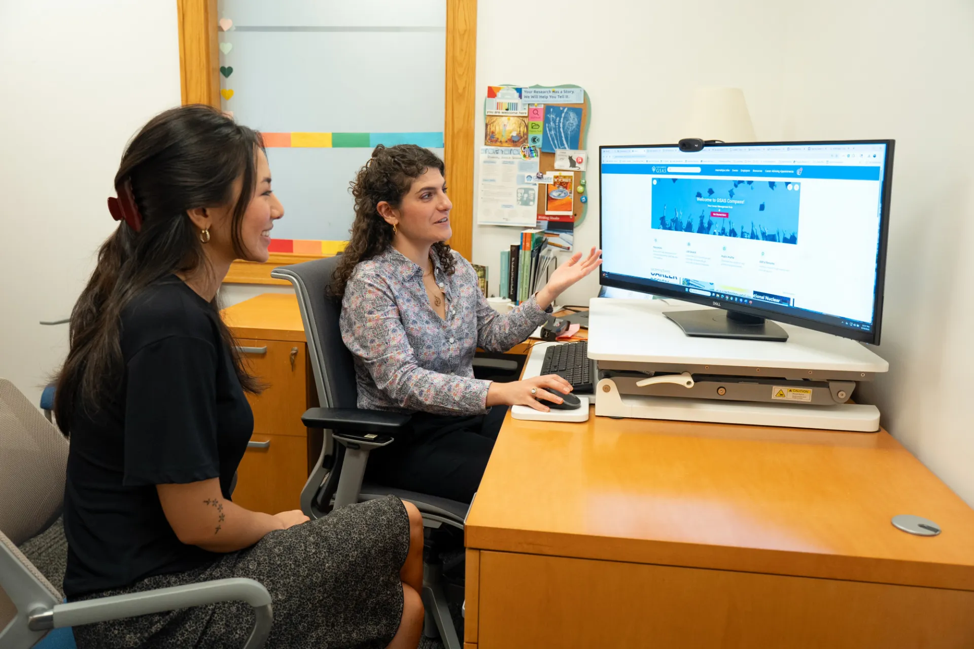 Two women in an office setting discuss content displayed on a large computer monitor. The scene conveys collaboration and a professional atmosphere.