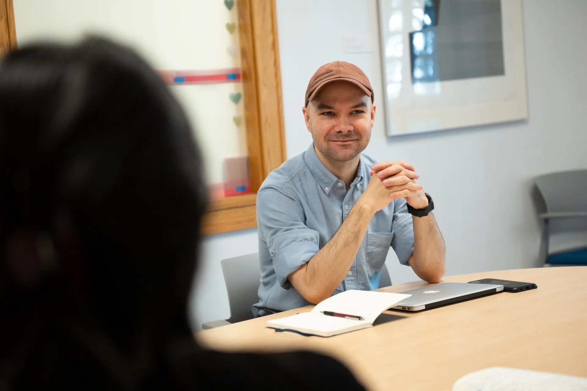A man in a casual shirt and cap sits at a table with a laptop and notebook, smiling warmly at a person in the foreground. The setting is relaxed and professional.