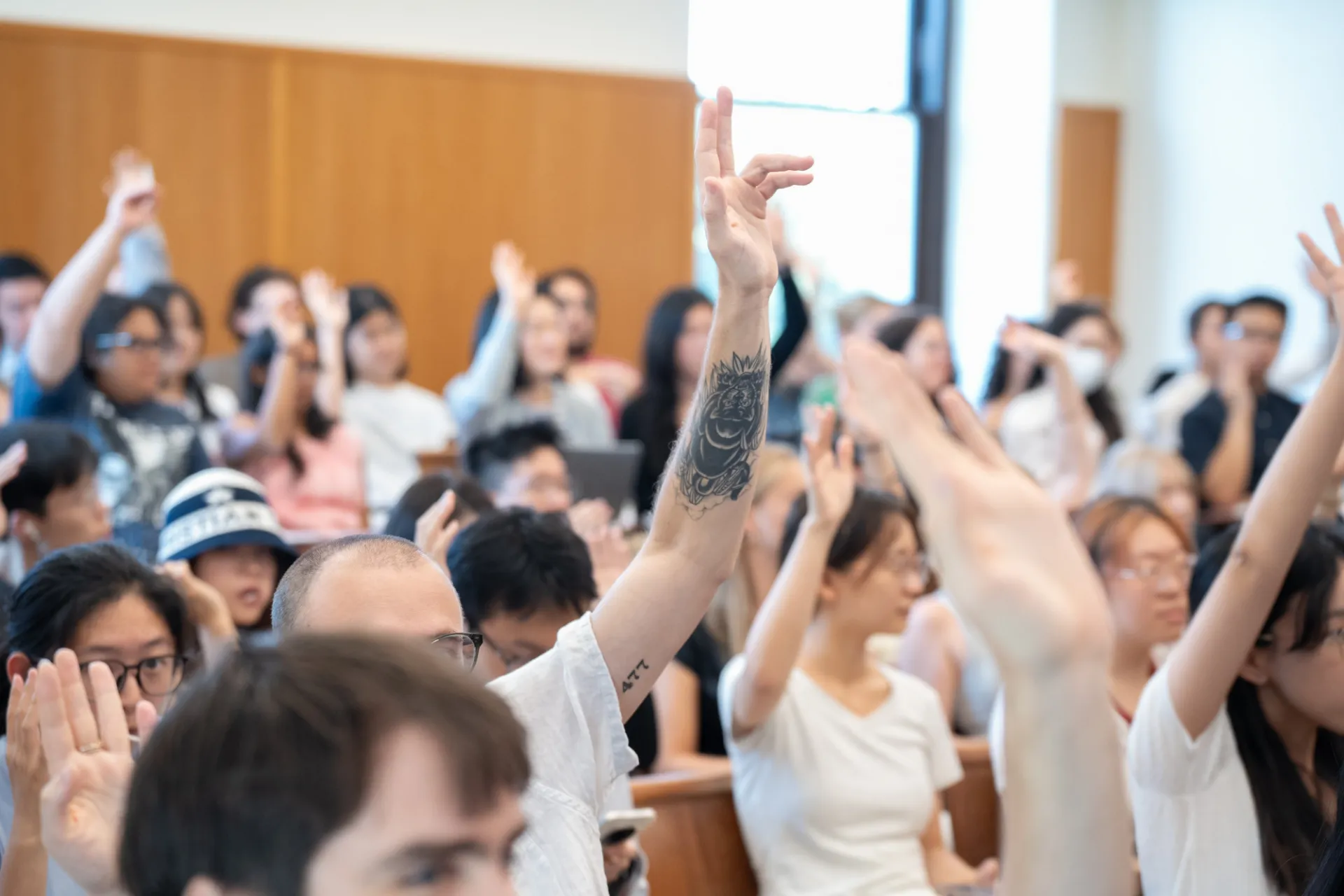 A classroom scene with diverse students seated, actively raising hands. Focus on a tattooed arm in the foreground, indicating engagement and curiosity.