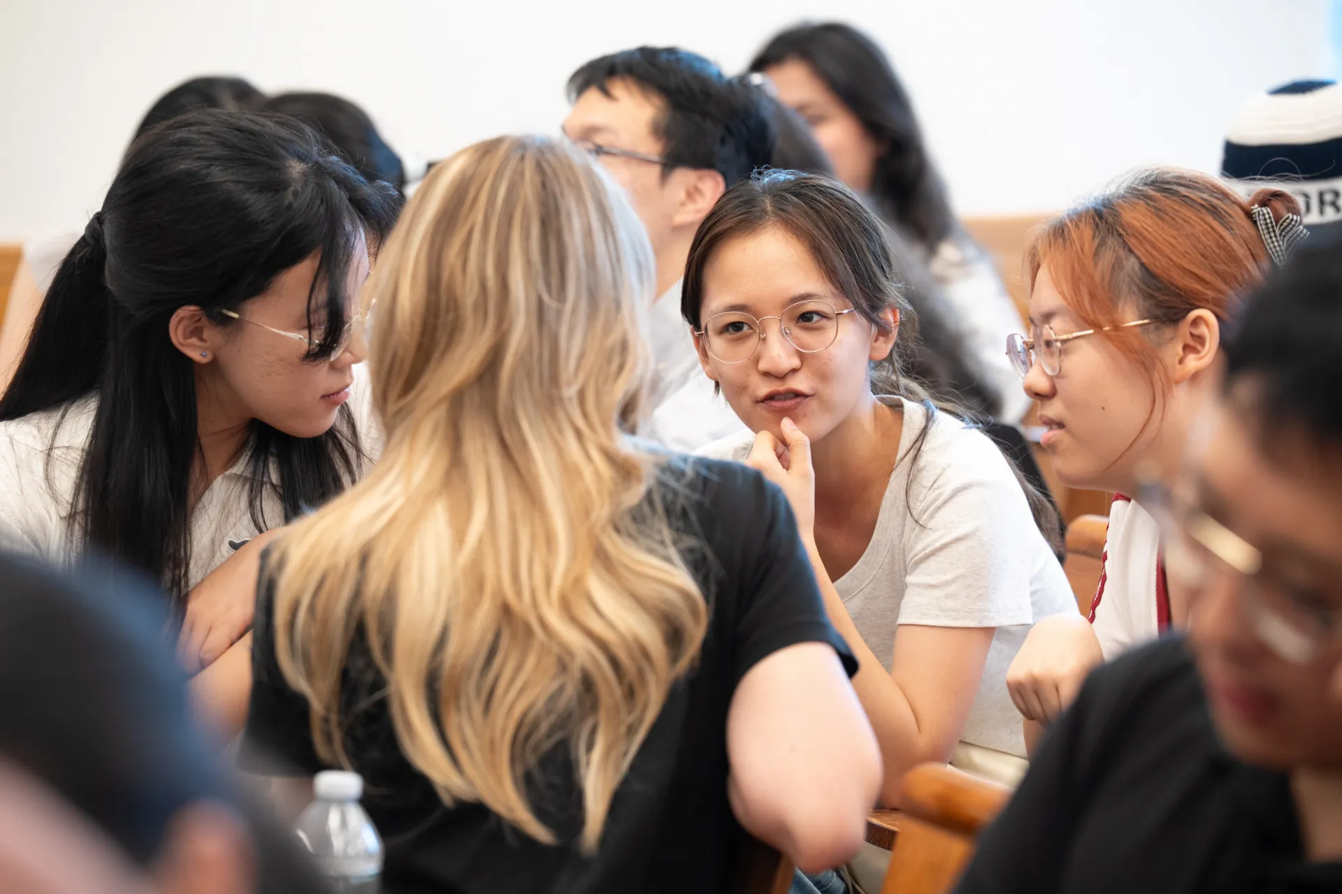 A diverse group of people engaged in animated discussion in a classroom setting. The atmosphere is focused and collaborative, with individuals leaning in to listen.