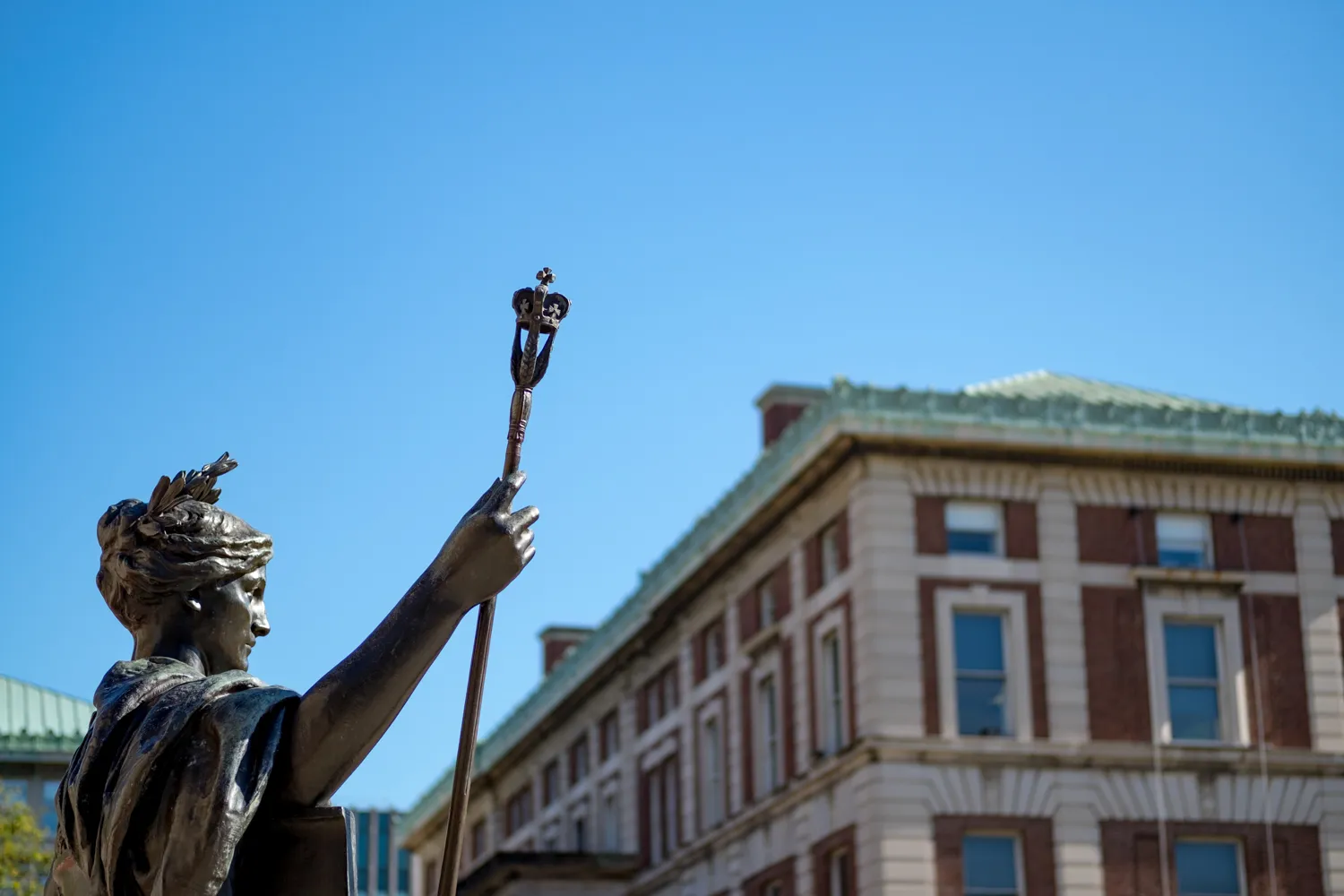 Statue of a robed figure holding a staff against a clear blue sky, with a classic brick building in the background. The scene conveys a sense of authority and history.