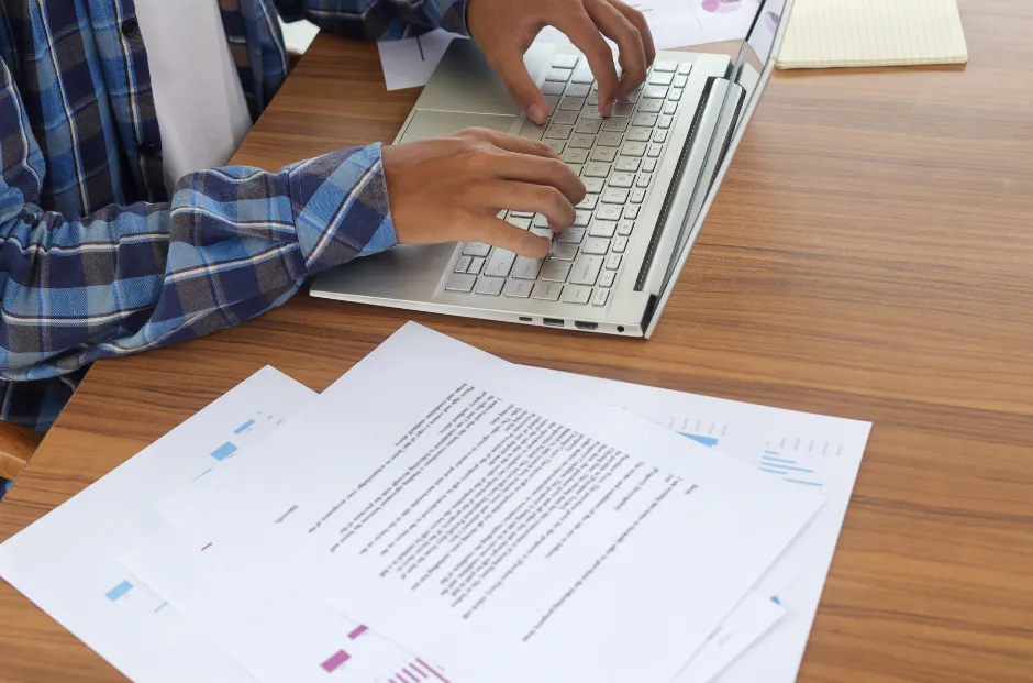A person in a blue plaid shirt types on a silver laptop at a wooden desk, surrounded by documents and graphs, creating a focused work atmosphere.