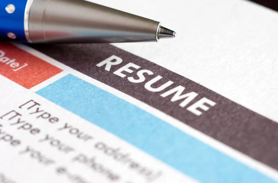 Close-up of a resume with bold text showing "RESUME" and a pen pointing to it. The page has colored sections, symbolizing organization.