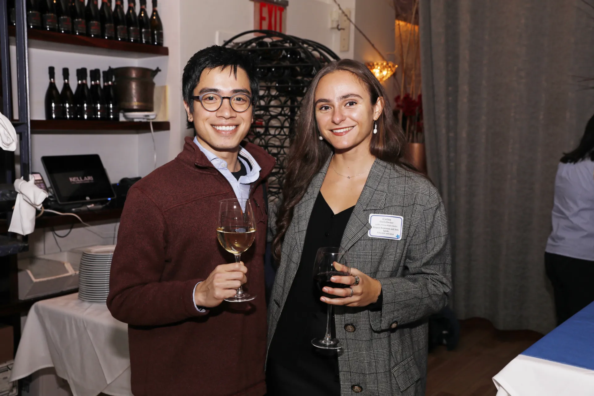 Two smiling people dressed professionally hold wine glasses at a networking event.