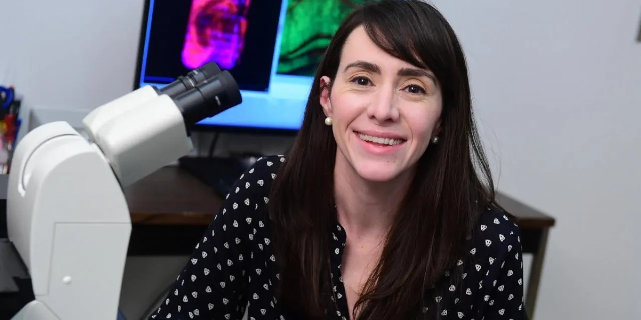 A woman with long brown hair smiles next to a microscope.