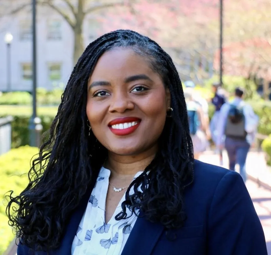 Smiling woman with braided hair and red lipstick stands in a sunlit park, wearing a dark blazer and patterned blouse. Trees and people in the background.