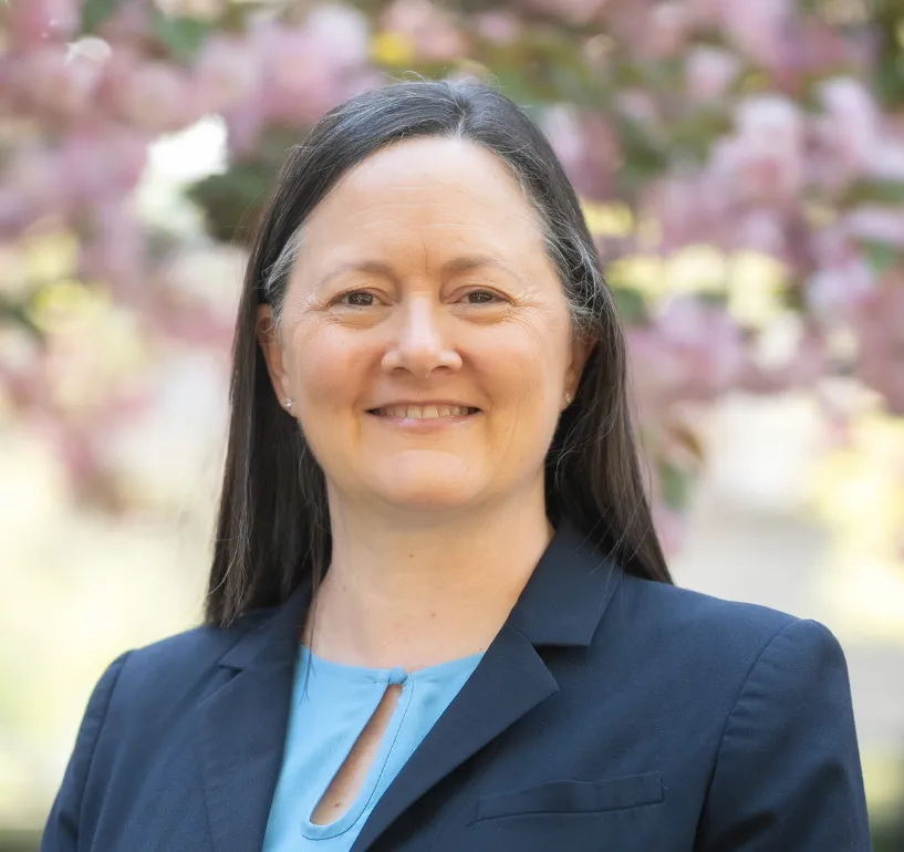 Smiling woman in a blue blouse and dark blazer stands outdoors. Pink blossoms and soft sunlight create a serene, pleasant background.
