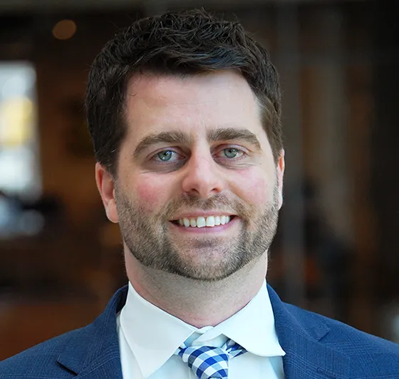 Smiling man in a blue suit and checkered tie stands in a warmly lit indoor setting. 