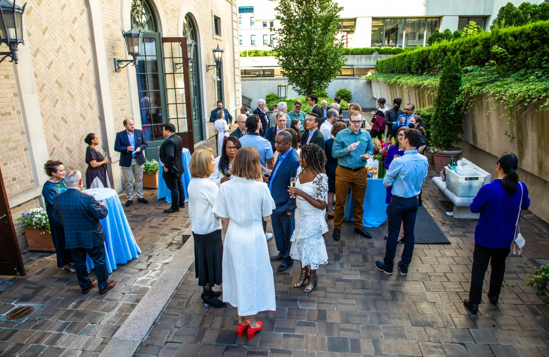 A group of people engage in lively conversation at an outdoor networking event. They stand on a brick patio, surrounded by greenery, exuding a vibrant and social atmosphere.