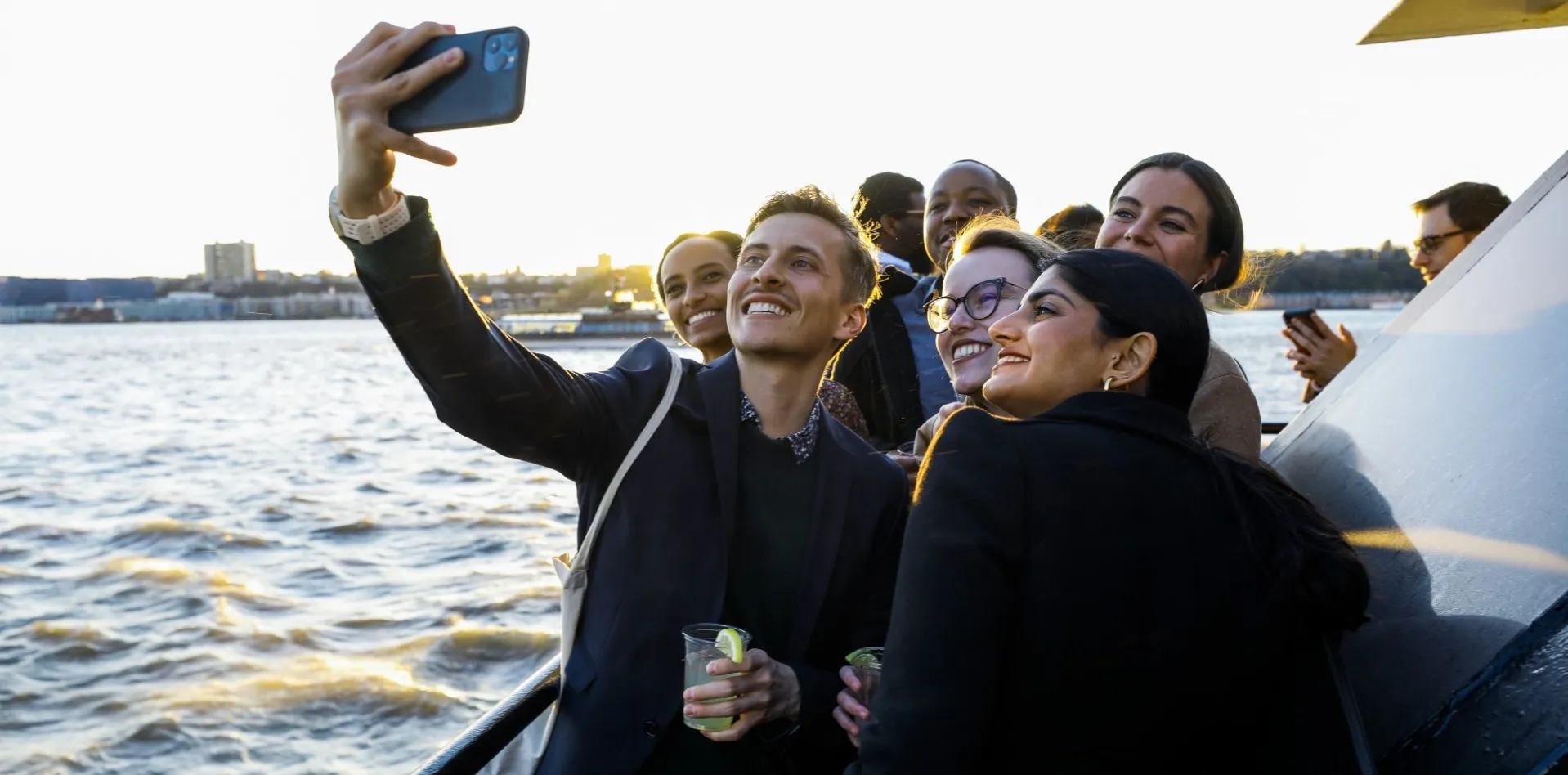 A group of people on a boat, smiling and taking a selfie against a sunset-lit river backdrop. The mood is joyful and celebratory.