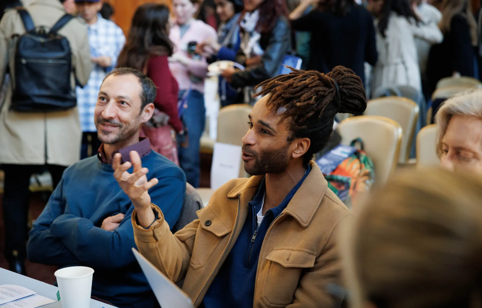 Two people engaged in conversation at a busy networking event. One gestures expressively; the atmosphere is lively and warm, with attendees in the background.