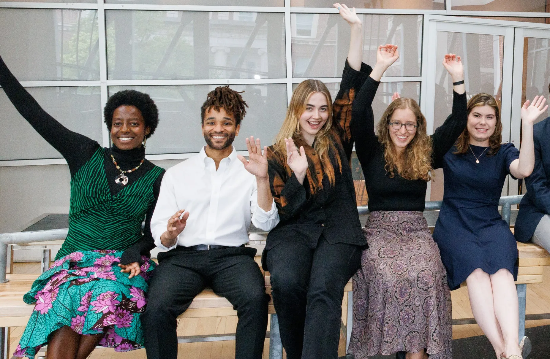 A group of people sit on a bench in a modern interior, joyfully raising their arms. They're dressed in smart-casual attire, conveying happiness.