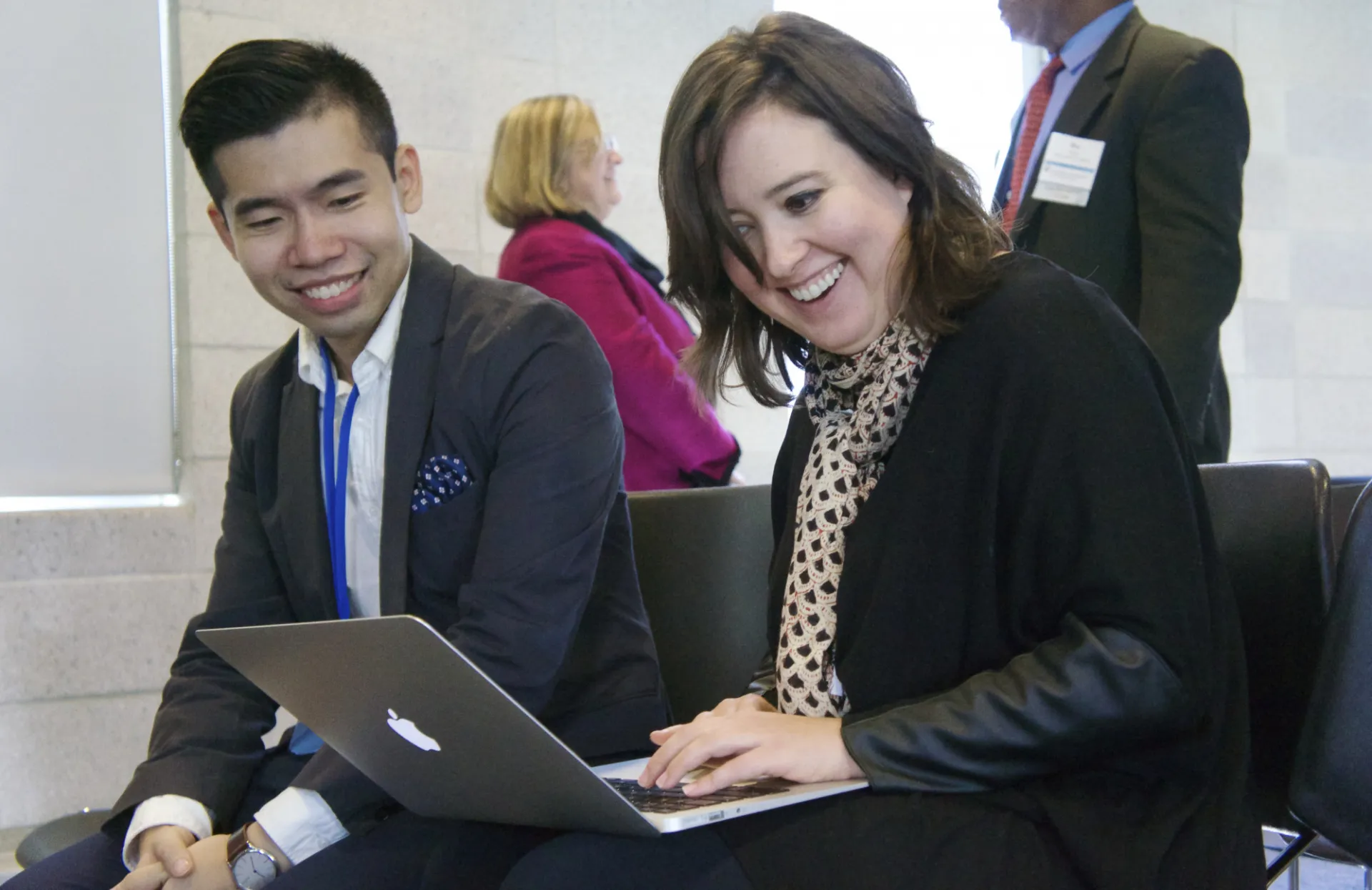 Two people sit closely, smiling and looking at a laptop. A woman, typing with enthusiasm, wears a polka-dot scarf. A man in a suit smiles. Lively atmosphere.