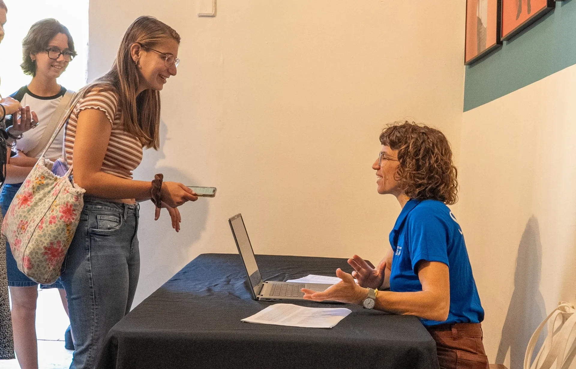 A woman in a blue shirt sits at a table with a laptop, assisting two people in line holding documents in a warmly lit room.