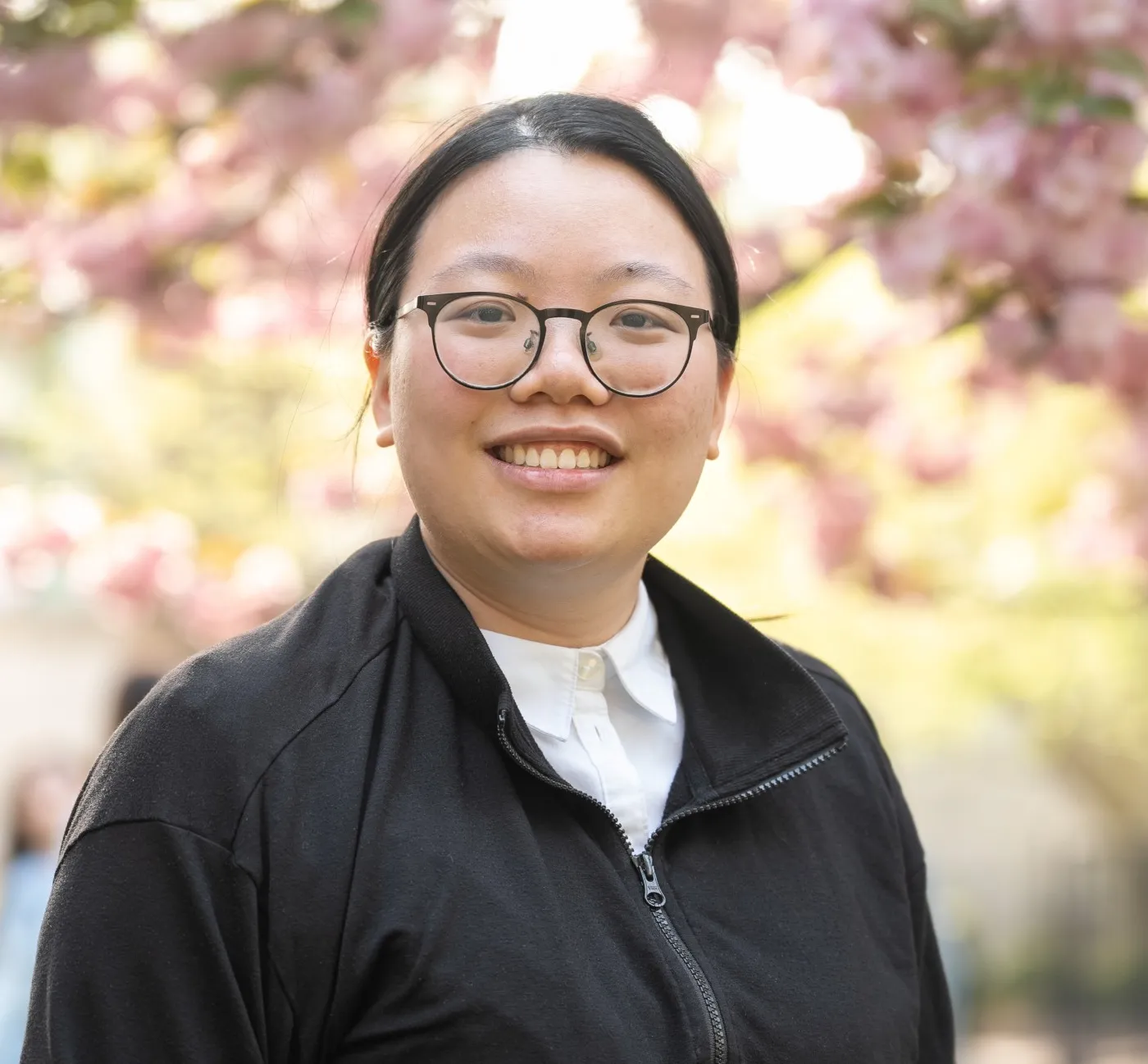 A woman with glasses smiling in front of a flowering tree.