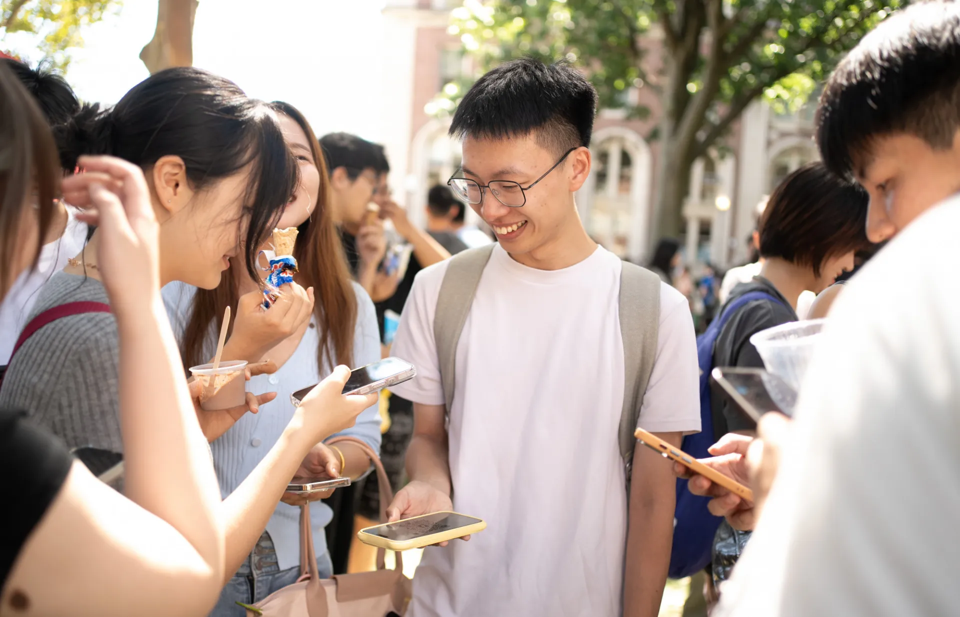 A group of young adults smiling and interacting outdoors with their smartphones. They are casually dressed, enjoying a sunny day in a lively, social atmosphere.