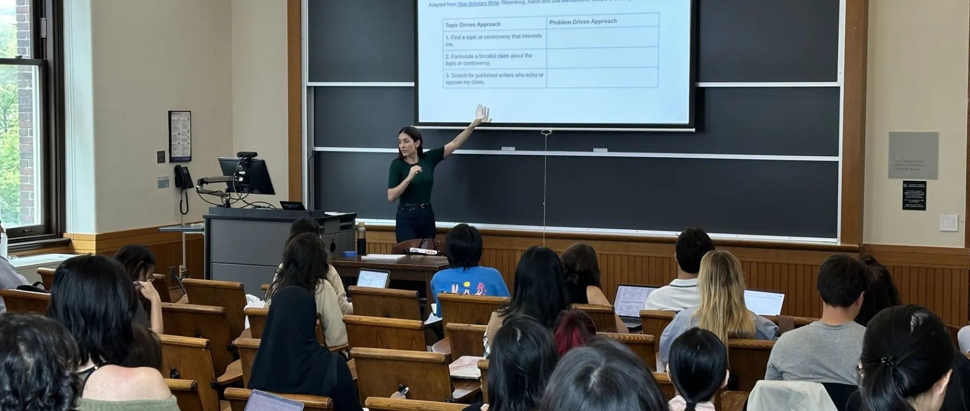 A lecturer in a classroom points to a projected slide titled "A Problem-Driven Writing Process." Students seated at desks focus on the presentation. The room has a studious atmosphere.