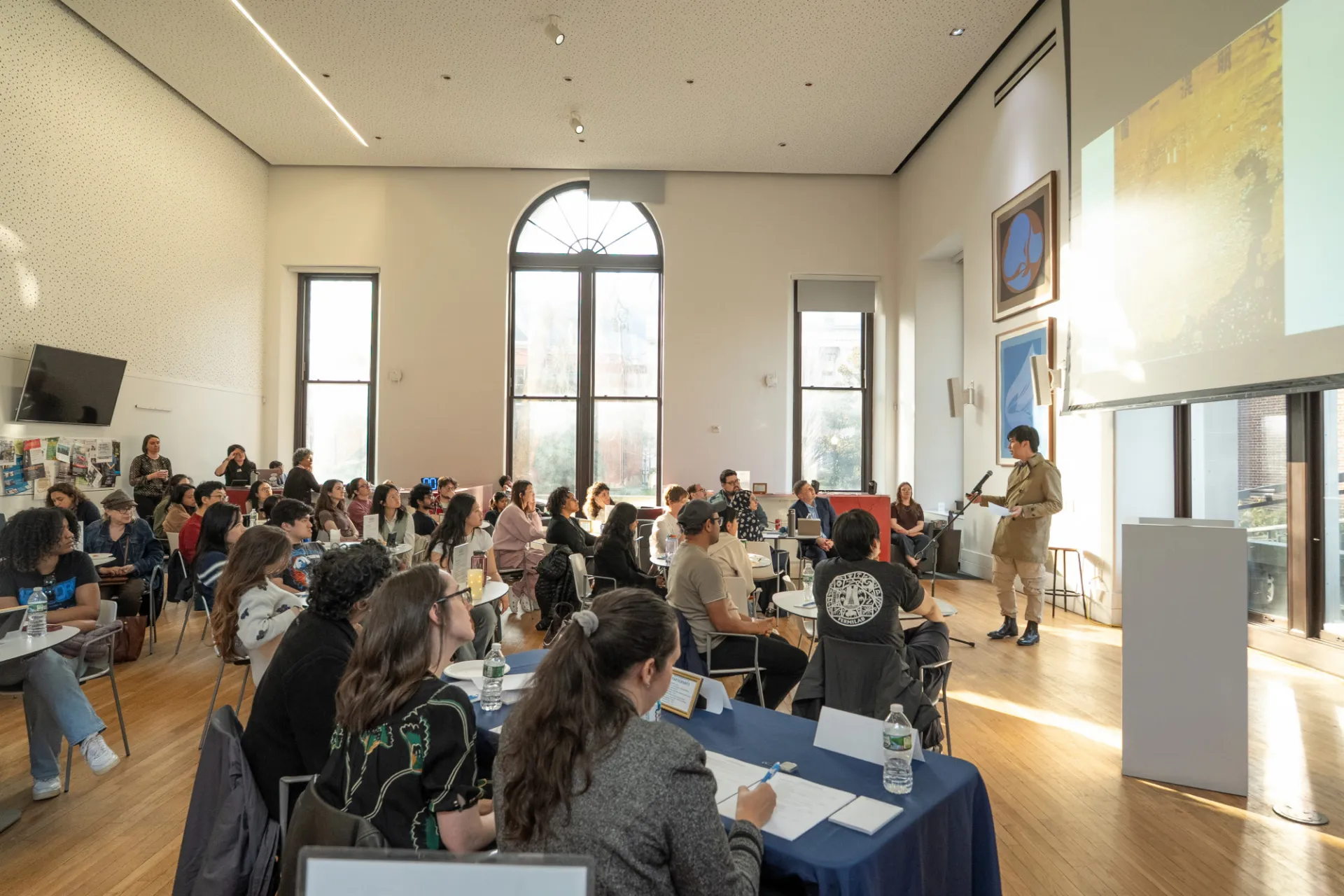 A wide shot of a man in a trench jacket with a projector screen behind him and students seated at tables listening to his presentation.