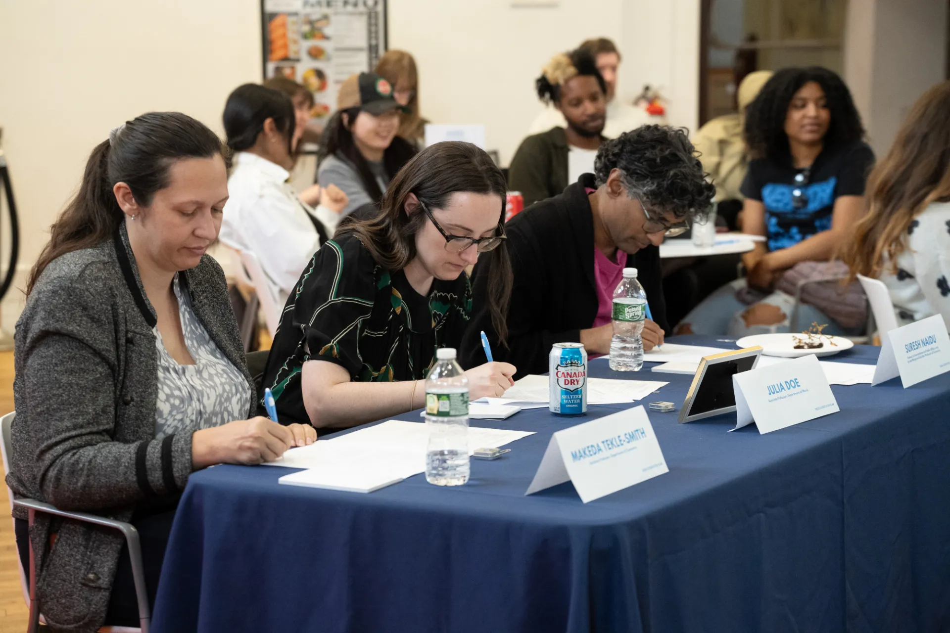 Three professors sit at a table writing down comments on their pieces of paper, with students seated in the background.