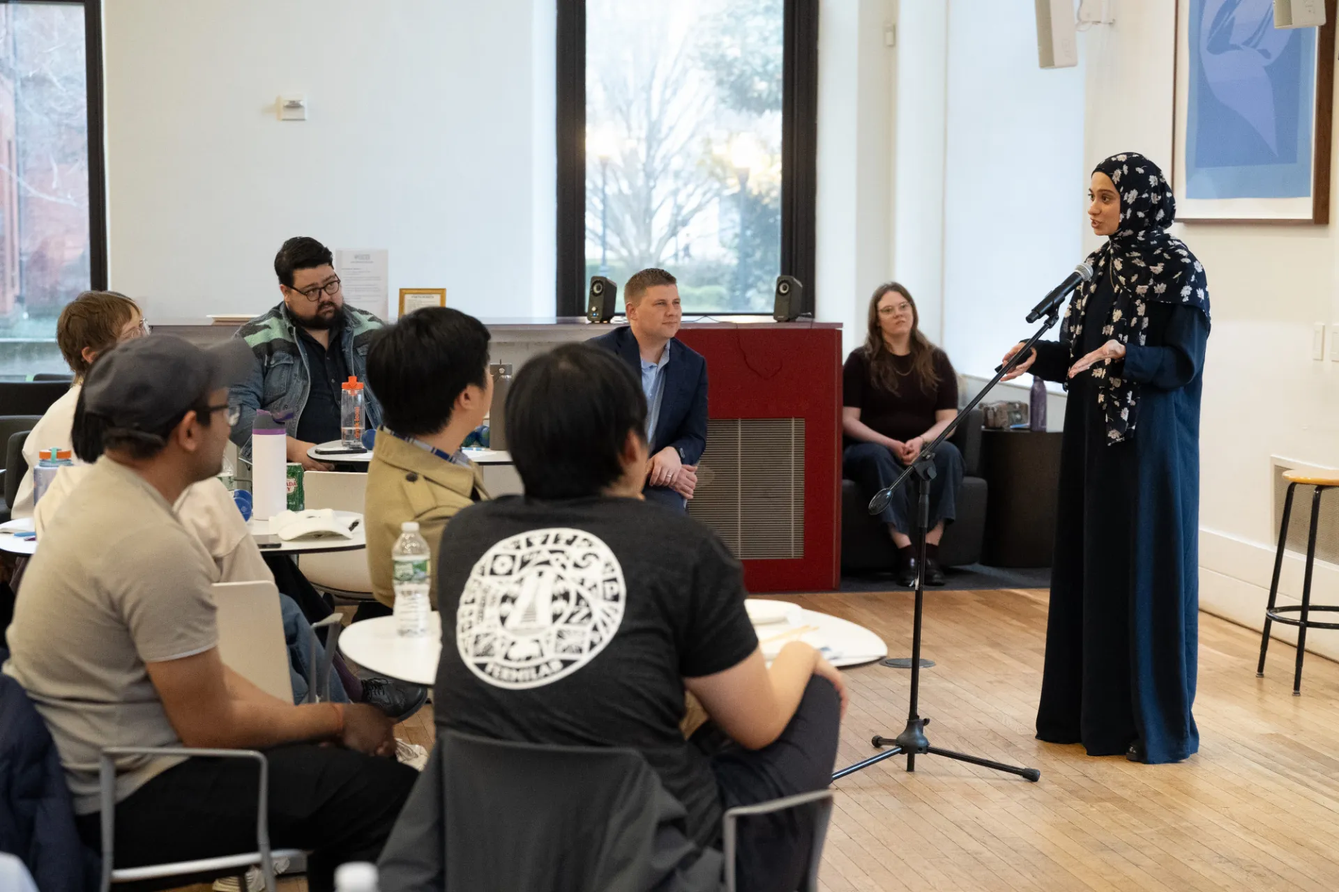 A woman wearing a floral-patterned hijab gives a speech at a mic stand in a light-filled room full of students sitting at tables listening attentively.
