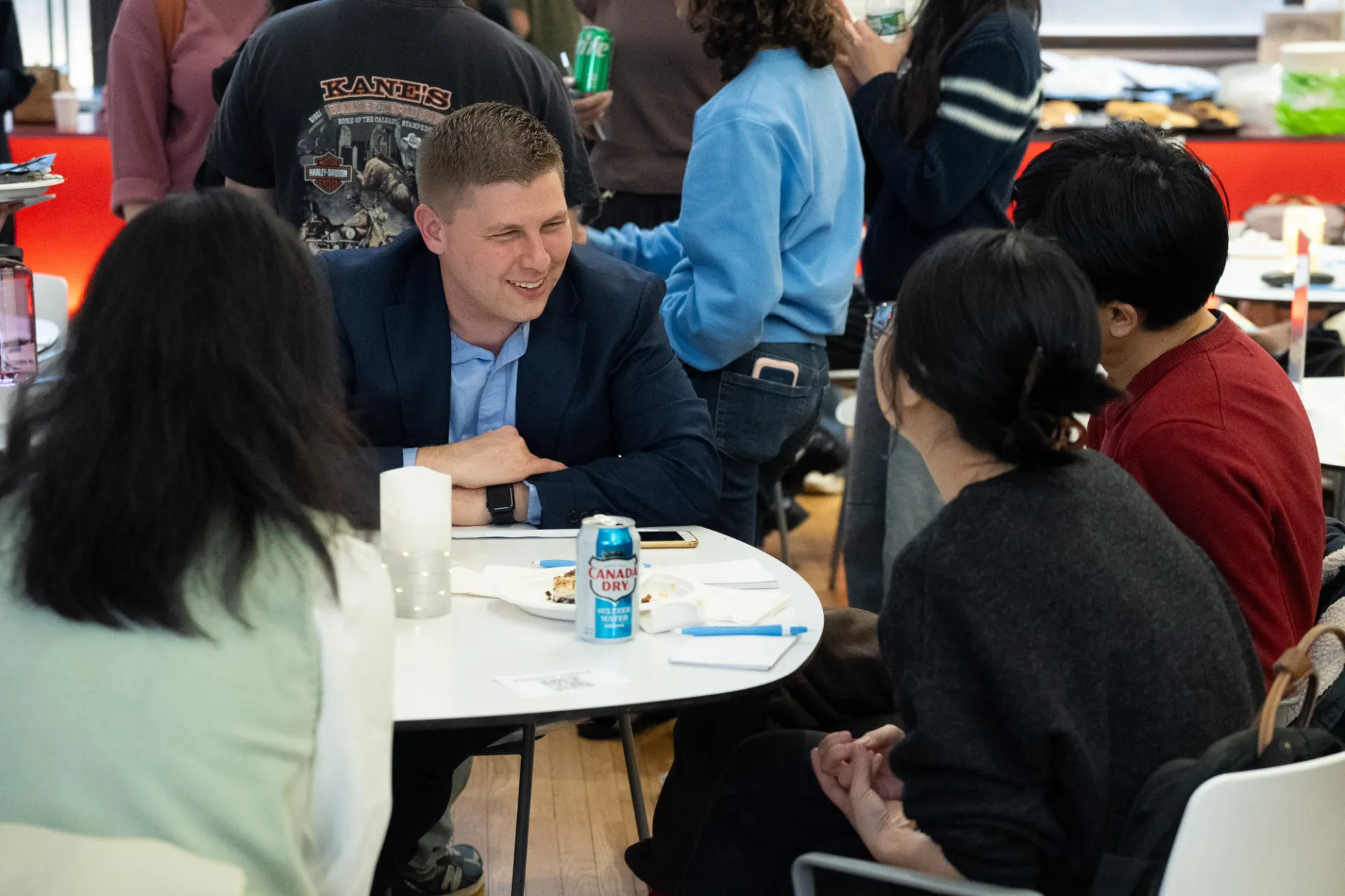 A man in a suit smiles at fellow students as they speak at a table.