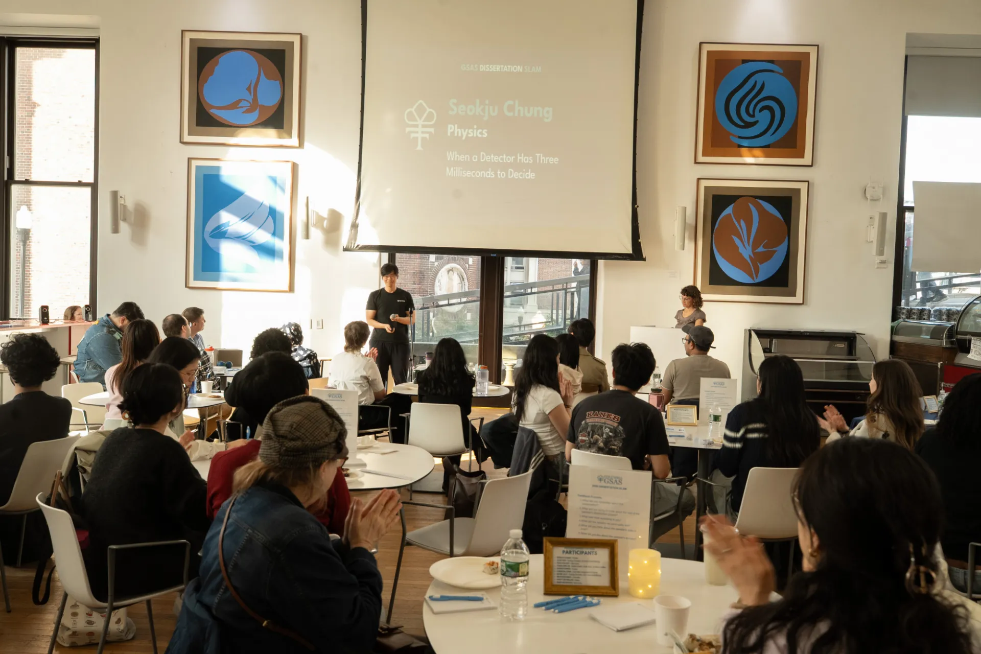 A man in a t-shirt and jeans with a projector behind him speaks in front of a room filled with students seated at white tables. 