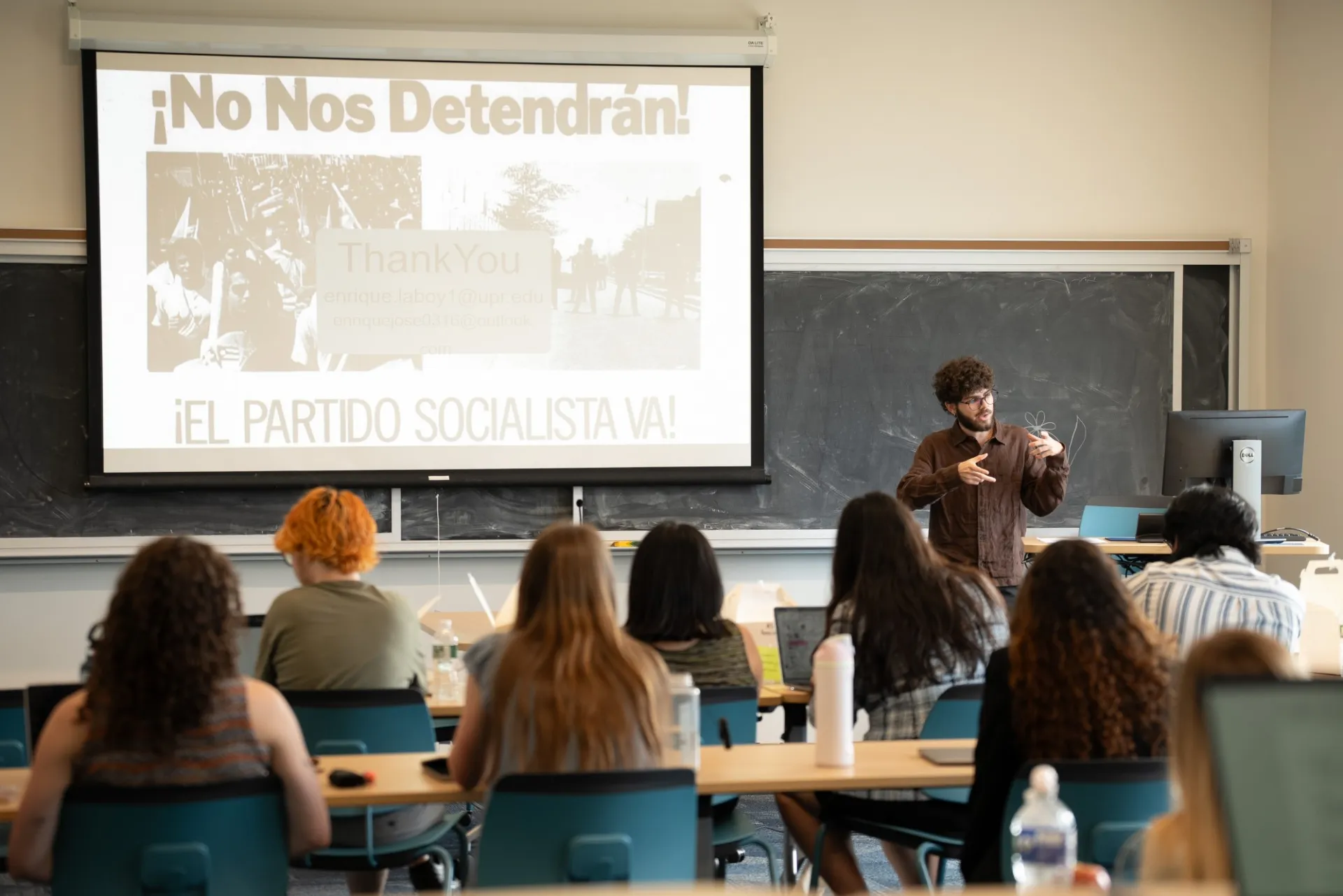 A person speaks passionately in front of a classroom. A presentation slide reads in Spanish, "¡No Nos Detendrán! ¡El Partido Socialista Va!" Students listen attentively.