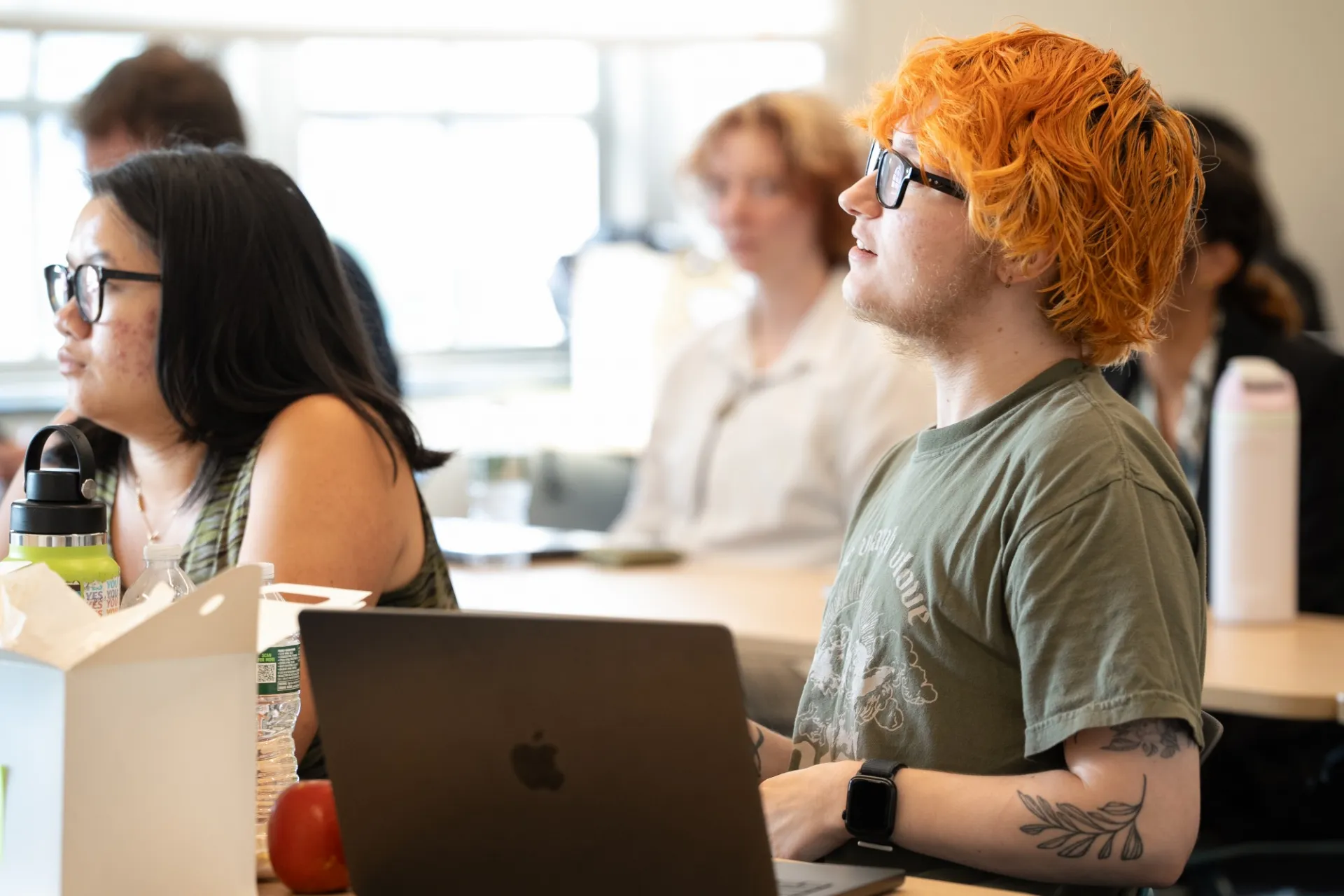 Students attentively listen in a classroom. A person with orange hair and tattoos faces forward near a laptop. The atmosphere seems focused and engaged.