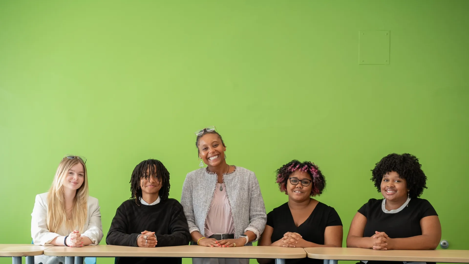 Five people, including a smiling woman standing behind a table, pose against a bright green wall. The mood is friendly and professional.
