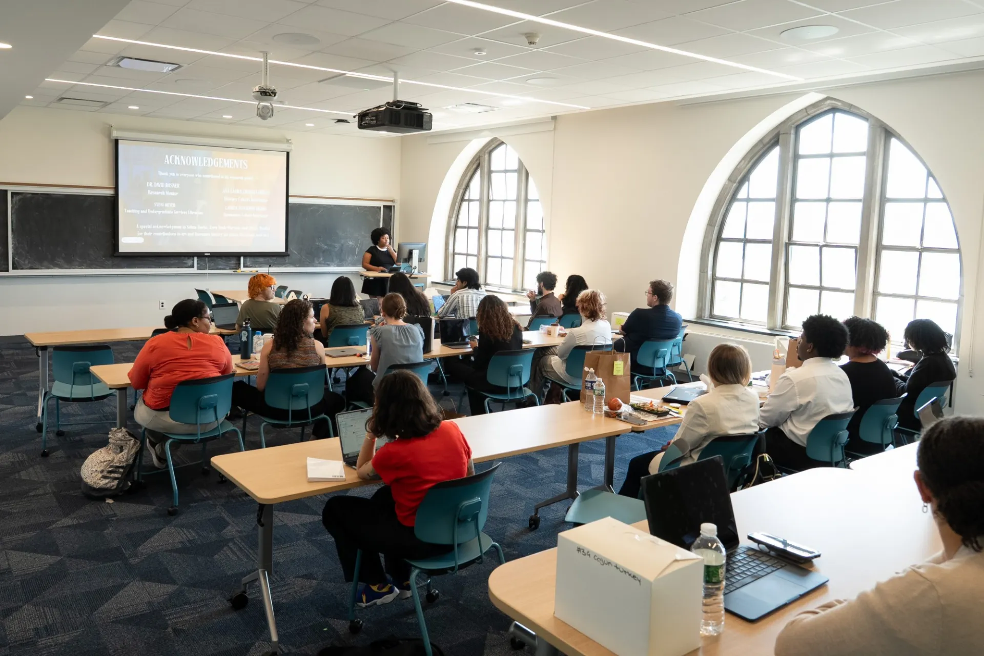 Classroom with curved windows, showing a diverse group of students sitting at tables listening to a presentation. A teacher stands at the front near a projected screen. Bright and focused atmosphere.