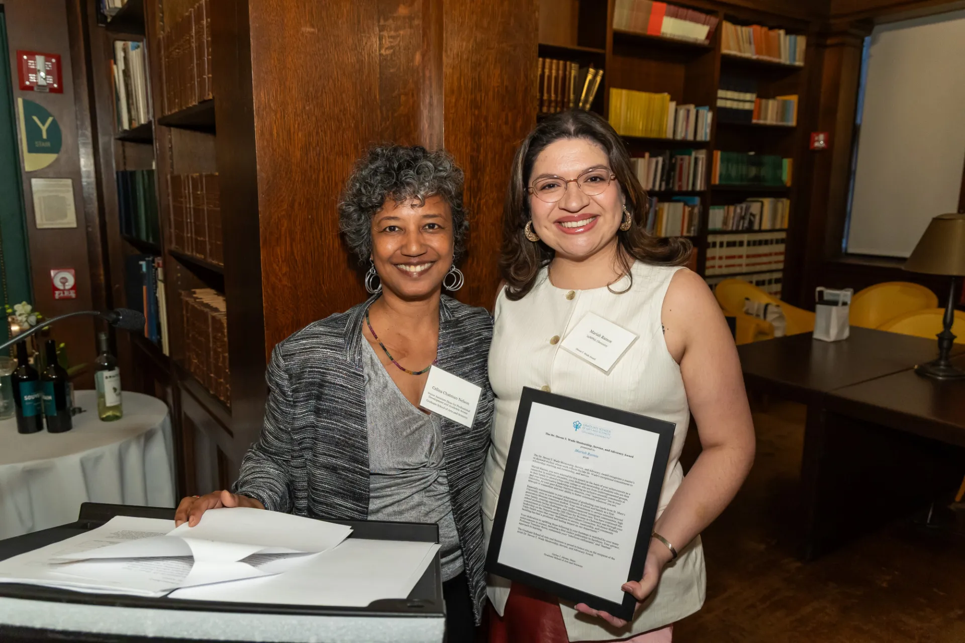 Two women dressed professionally, one with gray curly hair wearing a blazer, and one younger with brown hair and glasses, smile for a photo against a library backdrop while holding an award. 