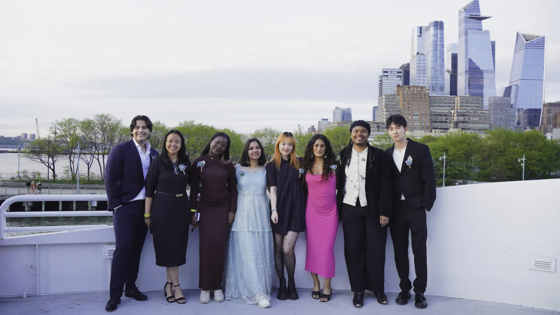 A group of formally dressed people in dresses and suits, pose for a group photo outdoors.