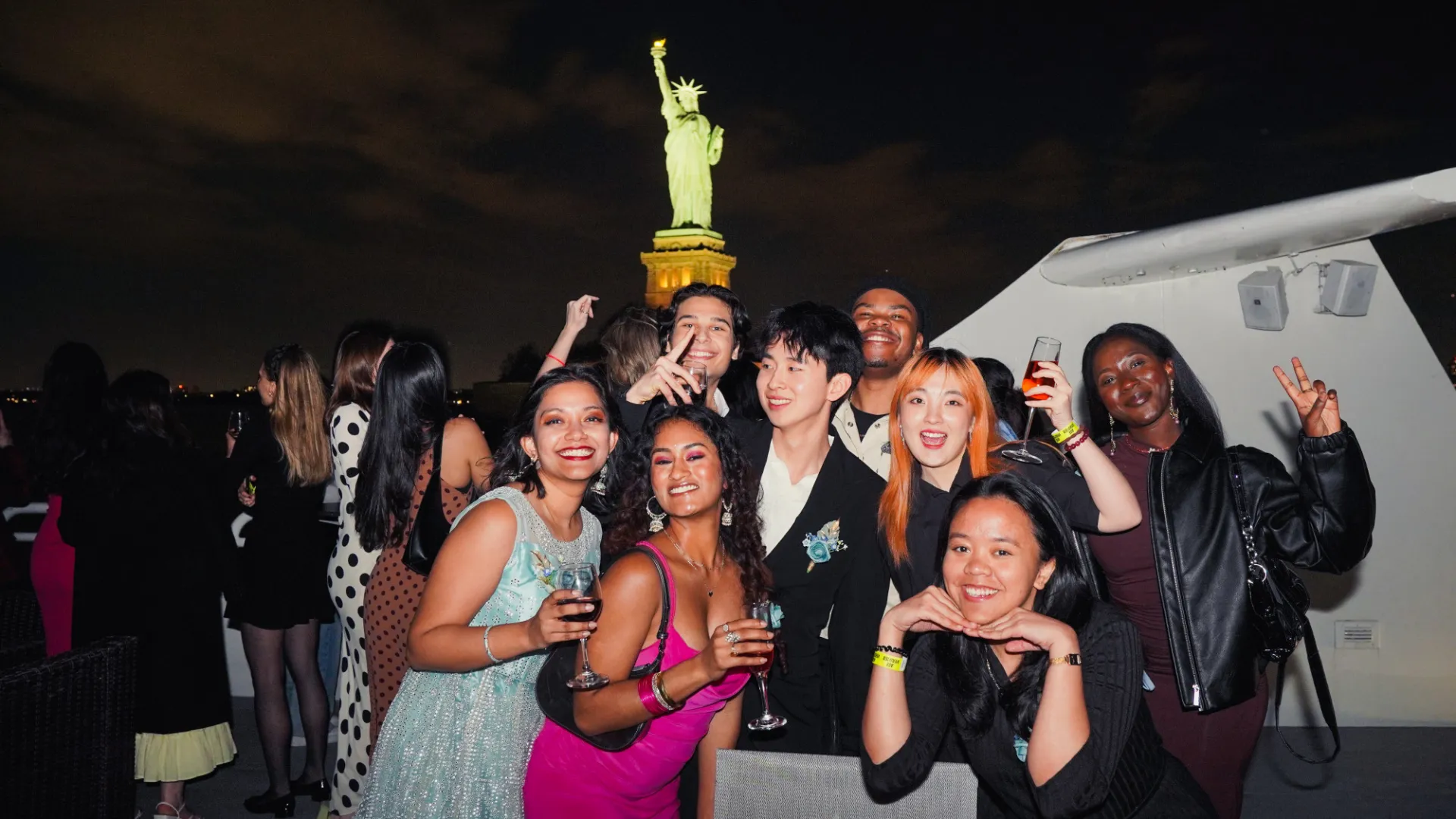 Students dressed festively holding wine glasses and smiling pose for a group photo at night on a boat, with the Statue of Liberty glowing in the background.