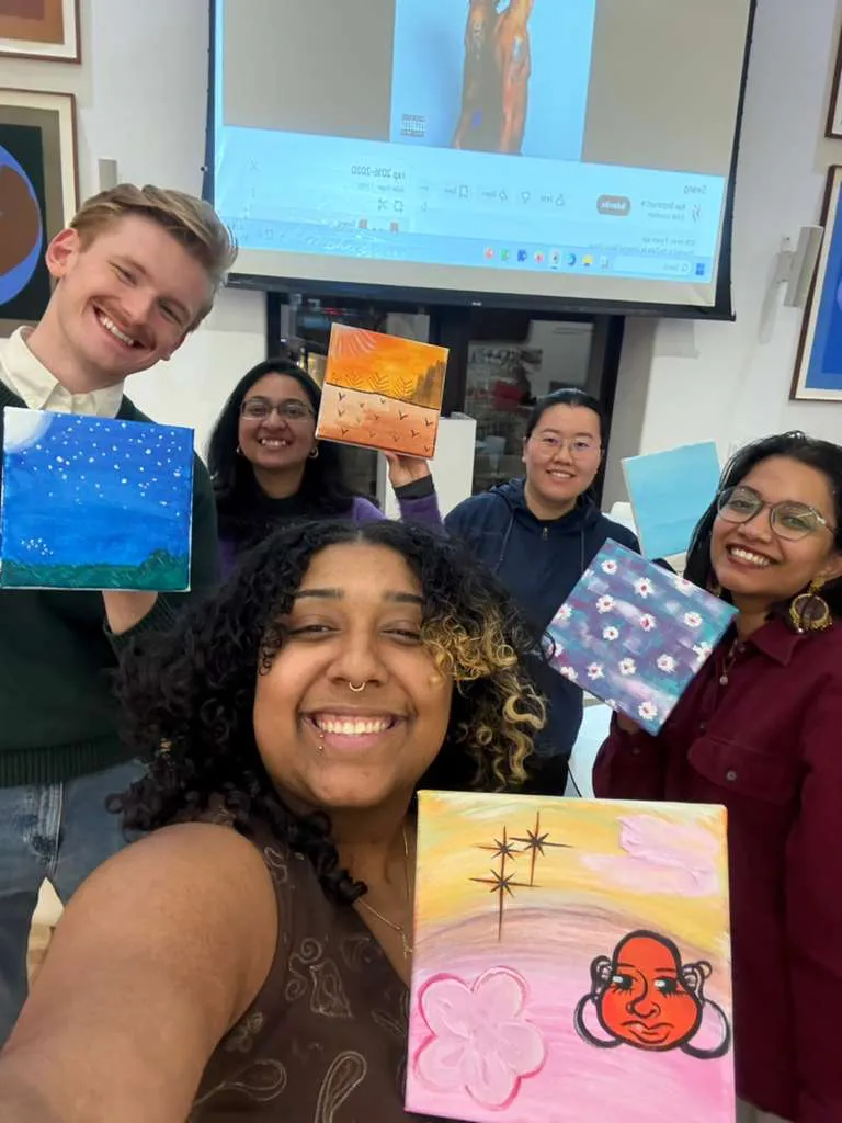 A selfie of smiling students holding small canvases with colorful paintings.