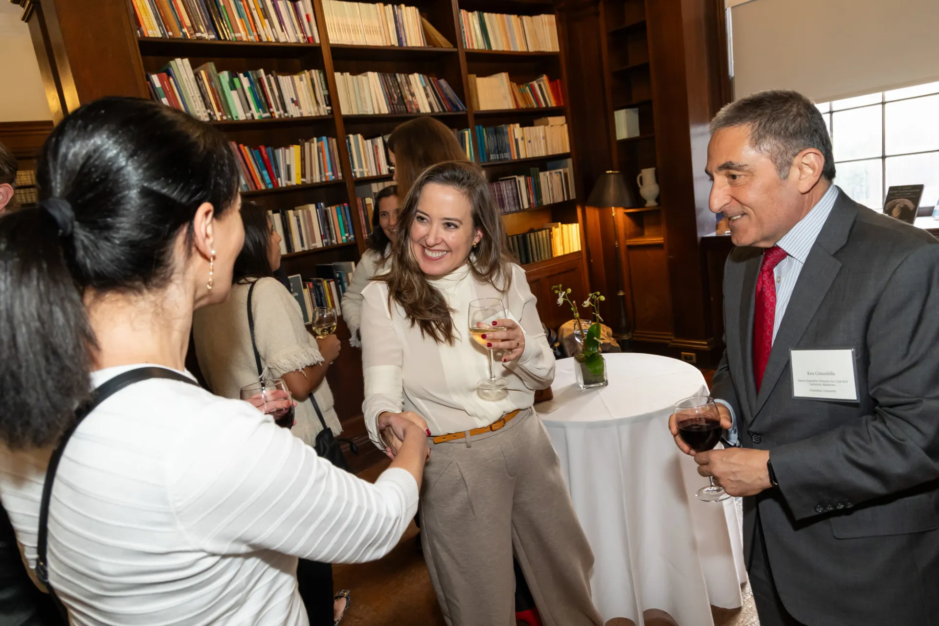 A woman dressed professionally holds a glass of wine and shakes someone else's hand at a reception.