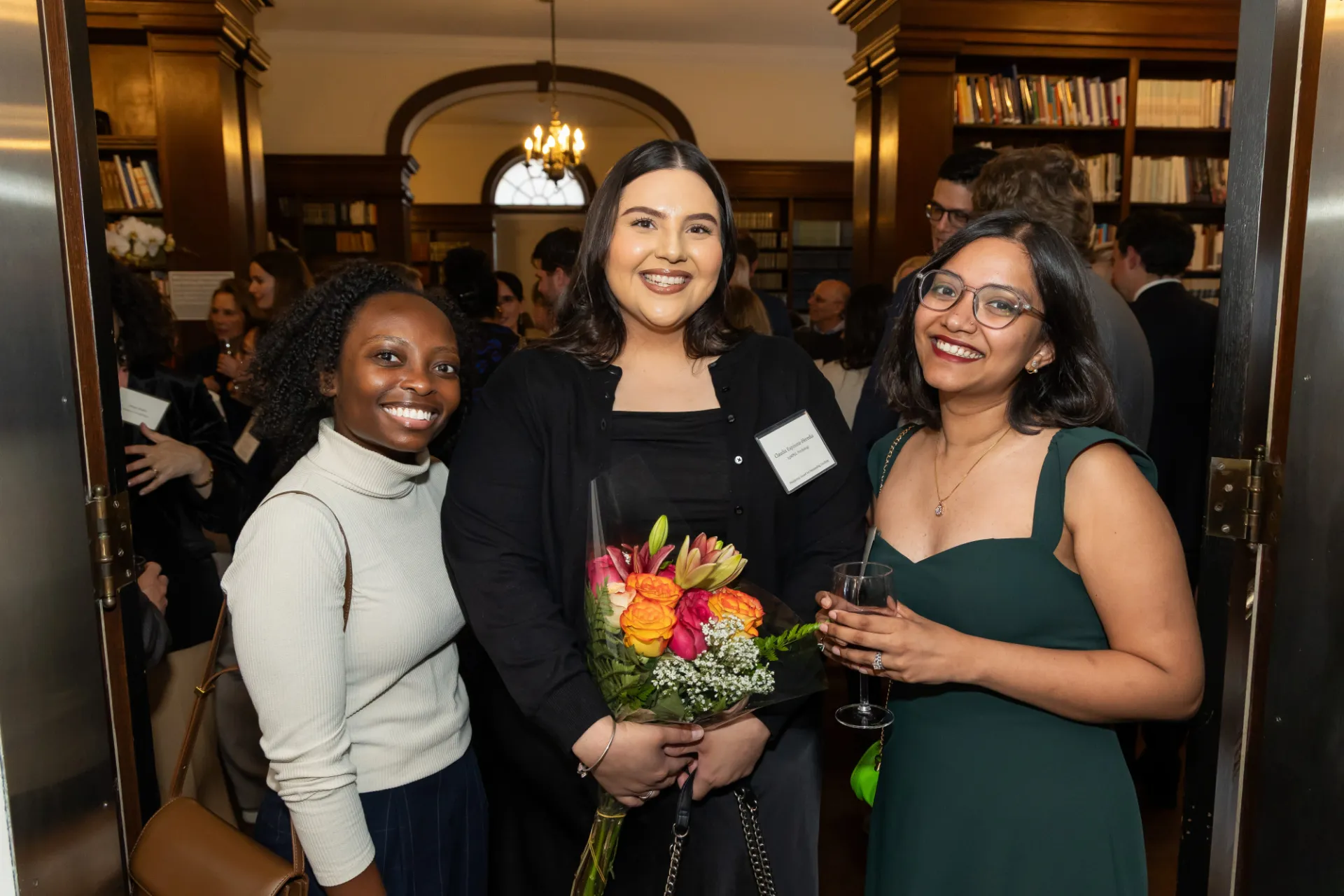 Three young women smile for a group photo, they are dressed for a reception, holding a bouquet of flowers.