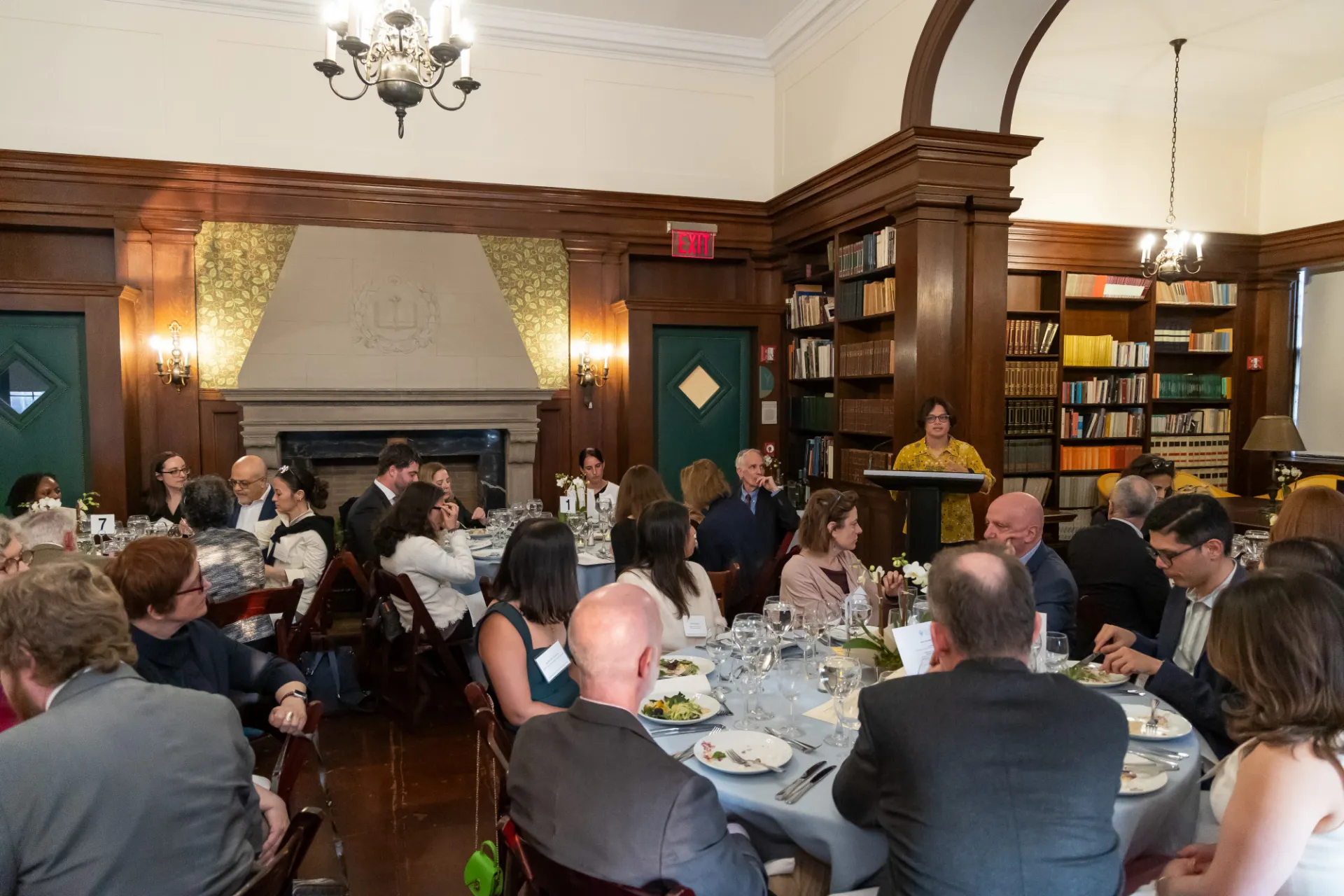 A woman stands at a podcium giving a speech to a wood-paneled room of diners seated at elegant tables. 