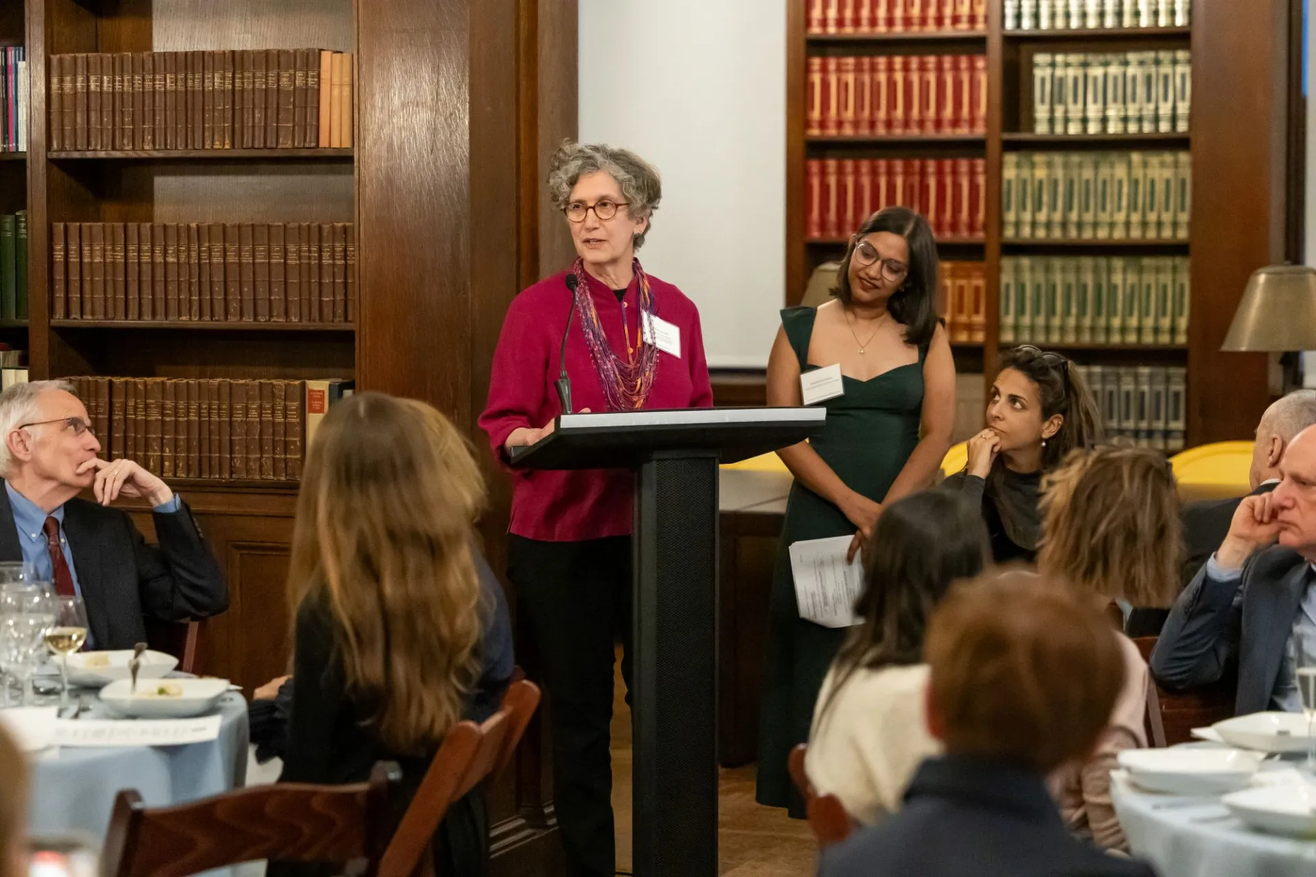 A woman with gray hair and glasses stands at a podium, speaking to a group of people seated at tables listening attentively. She is in a room lined with books. 
