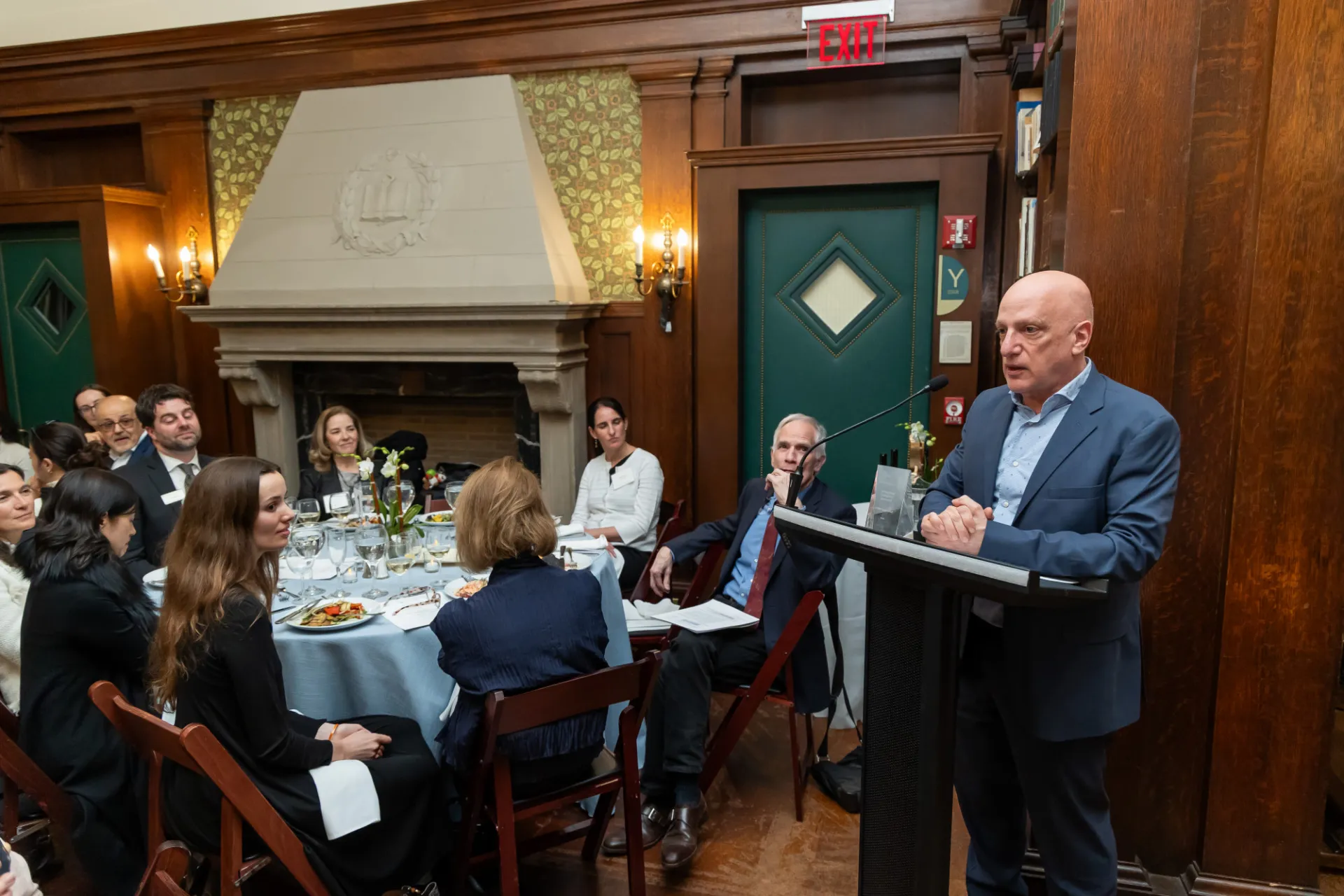 A man in a gray suit speaks seriously at a podium, attendees sit at tables with food, listening to his speech.