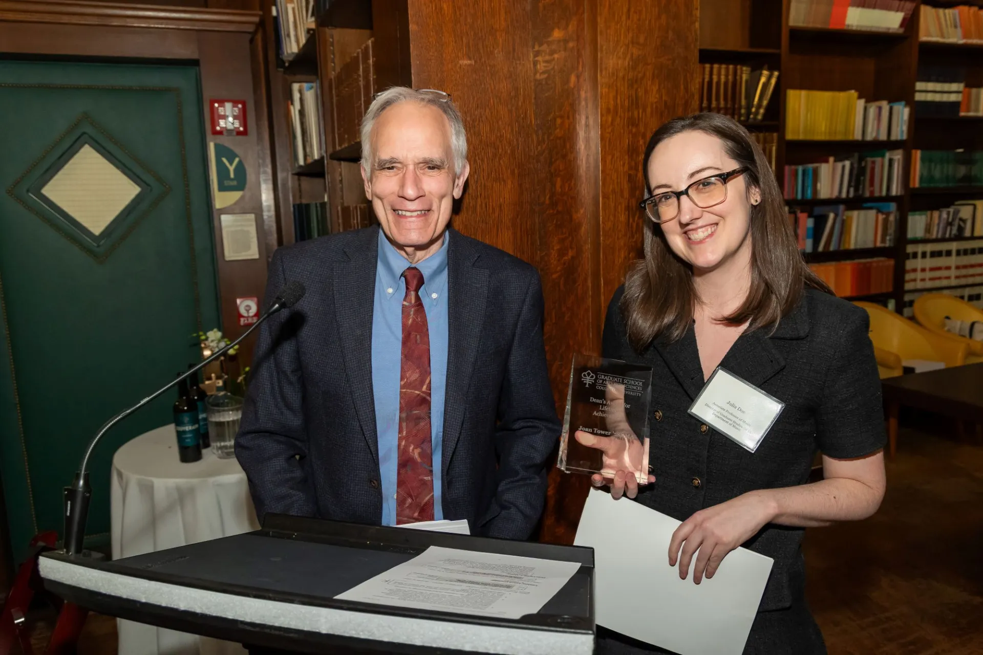 A woman with glasses in a black dress holds a crystal award and smiles at the podium with a man in a suit.