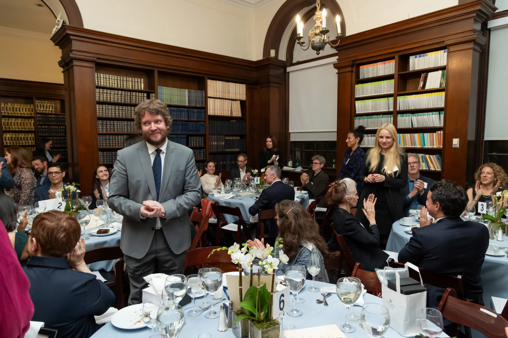 A man in a suit and a woman in a black suit stand at tables, while guests applaud them.