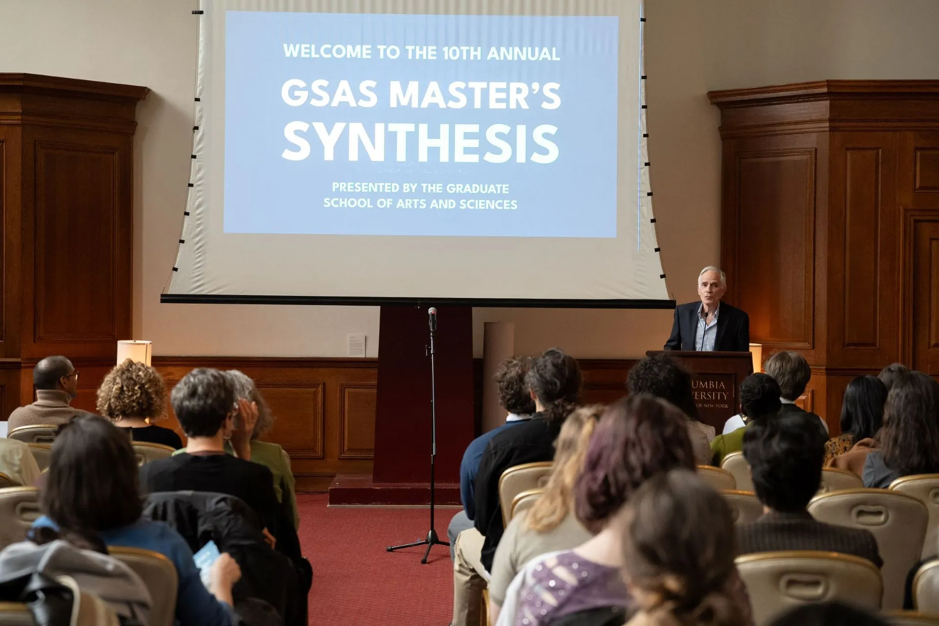 Dean Carlos speaks at a podium before a large audience. 
