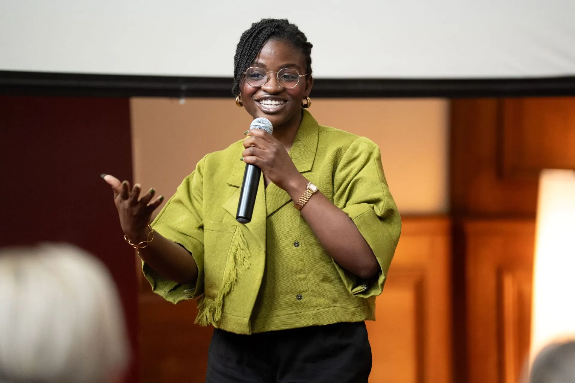 A woman in glasses wearing a green short-sleeved blazer smiles as she talks expressively into a microphone.